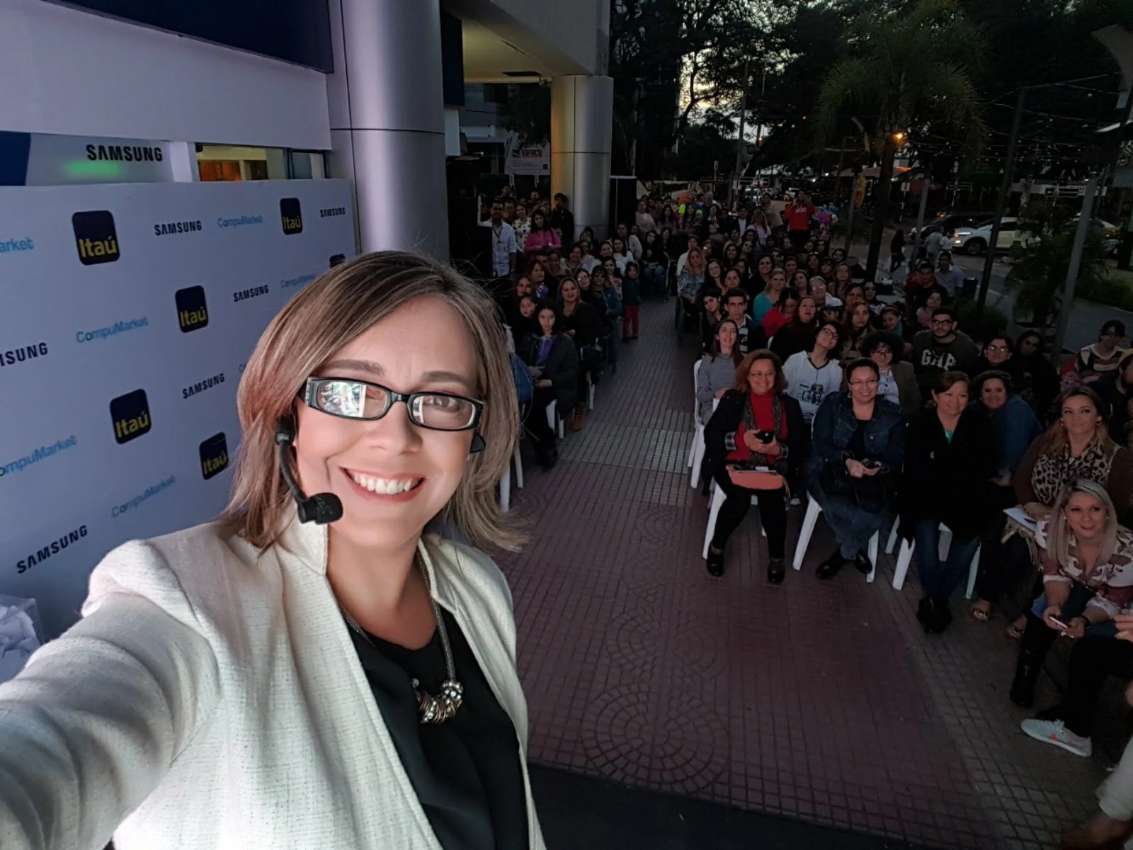 Una mujer con gafas y micrófono toma una selfie en un evento con muchas personas sentadas en sillas al aire libre, al fondo se ven árboles y más gente, en un entorno de noche.