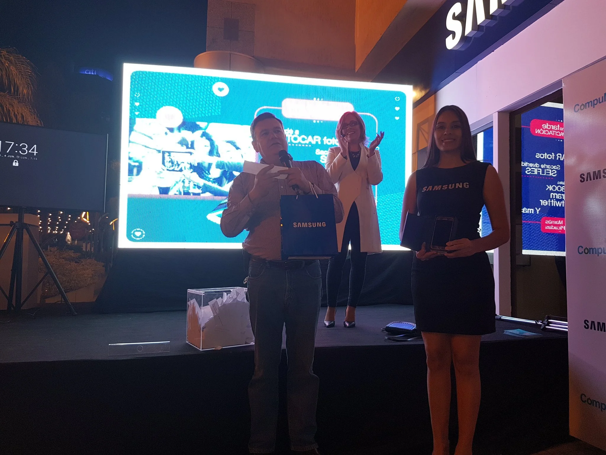 Personas en un evento de Samsung en un escenario con pantalla grande de fondo, una mujer con camiseta negra de Samsung en primer plano, un hombre con micrófono y bolsa, y otra mujer en el escenario, todos en un ambiente nocturno.