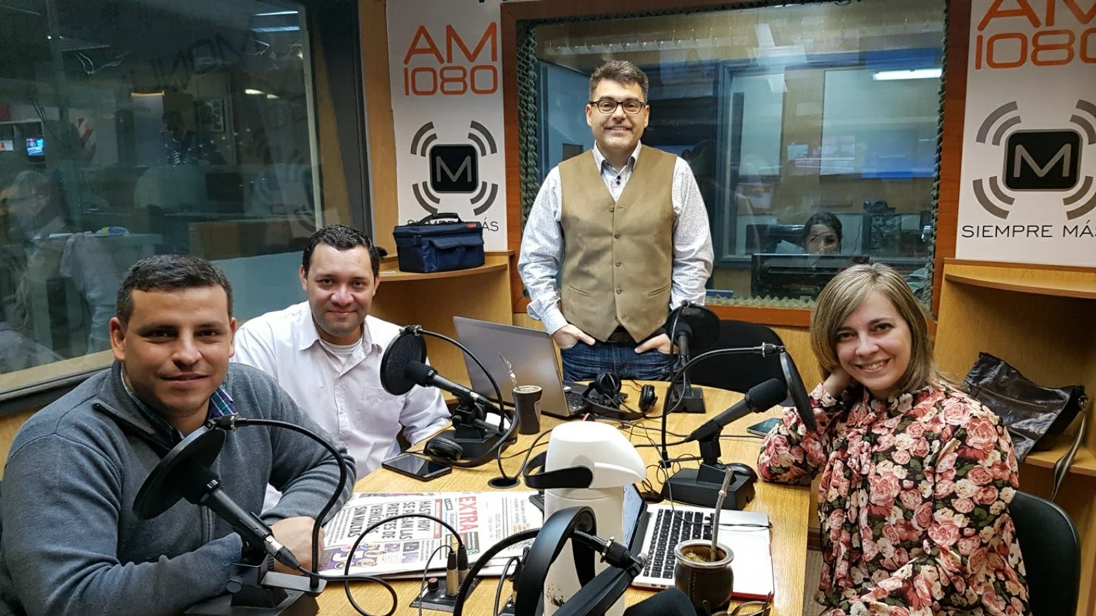 Cuatro personas en un estudio de radio con micrófonos, computadora, y periódicos, posando para la foto en un ambiente de transmisión en vivo.