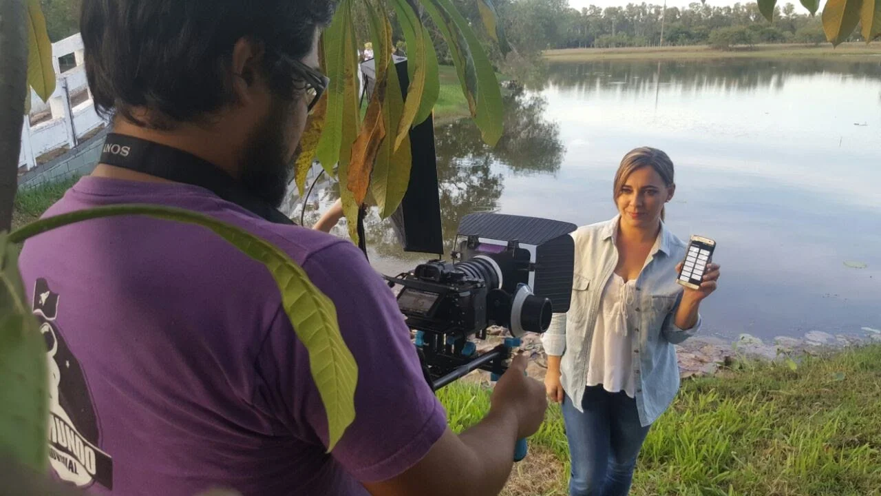 Una mujer posa frente a un lago mostrando su teléfono móvil con una aplicación en la pantalla, mientras un hombre con cámara la fotografía desde atrás, en un entorno natural con árboles y agua.