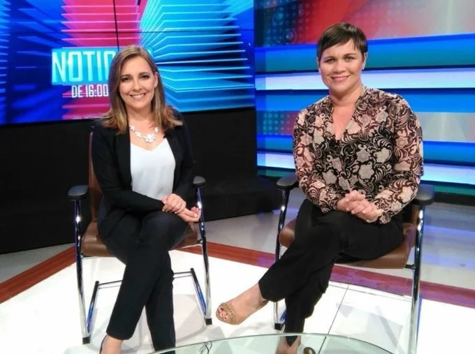 Dos mujeres sentadas en un estudio de televisión, con pantallas de fondo y un letrero que dice 'NOTICIAS'.