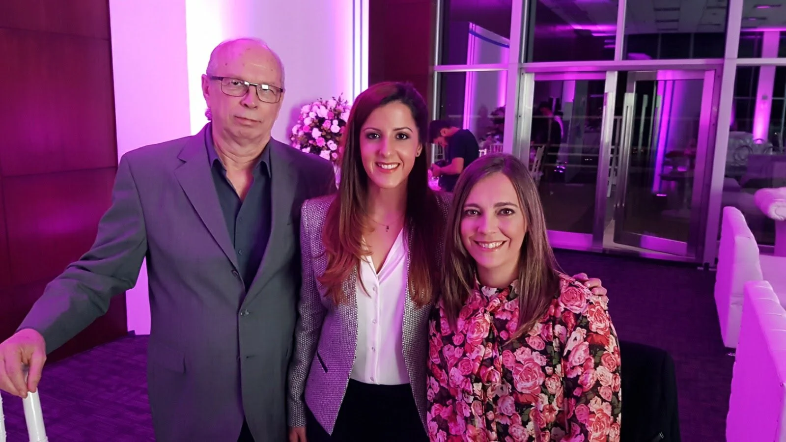 Tres personas en una fiesta con iluminación morada, sonriendo y posando para la foto, en un recinto con fondo de vidrio y flores.