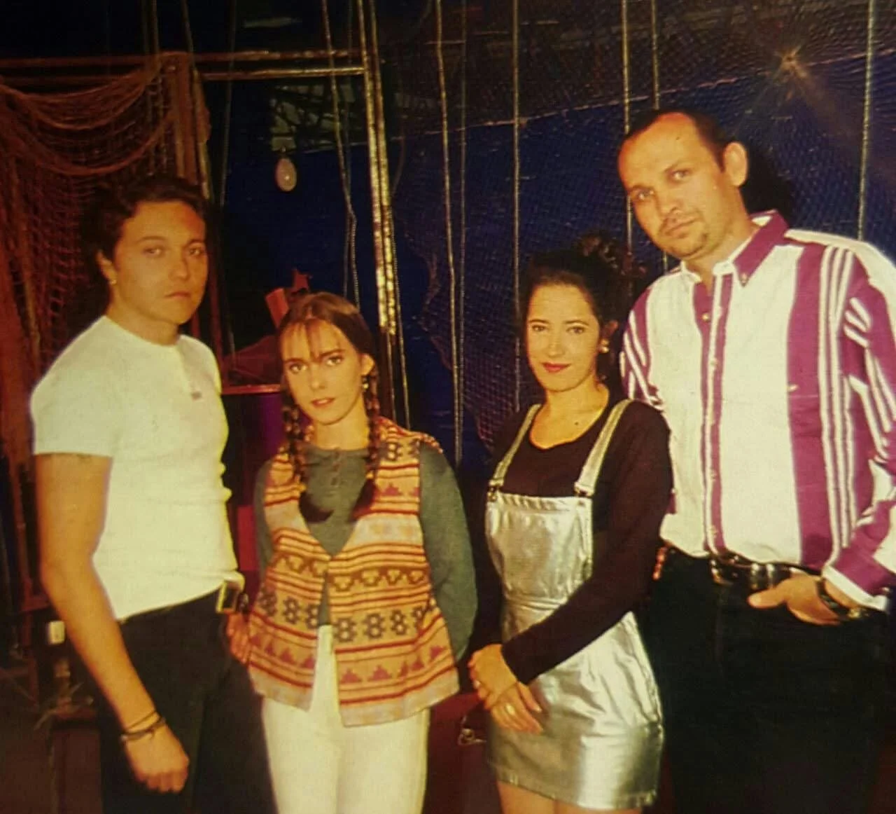 Cuatro personas en un escenario con decoraciones de circo o teatro, incluyendo telarañas y cortinas, una mujer con vestimenta en colores oscuros y dos con ropa clara, y un hombre con camisa de rayas moradas, blancas y rosas.