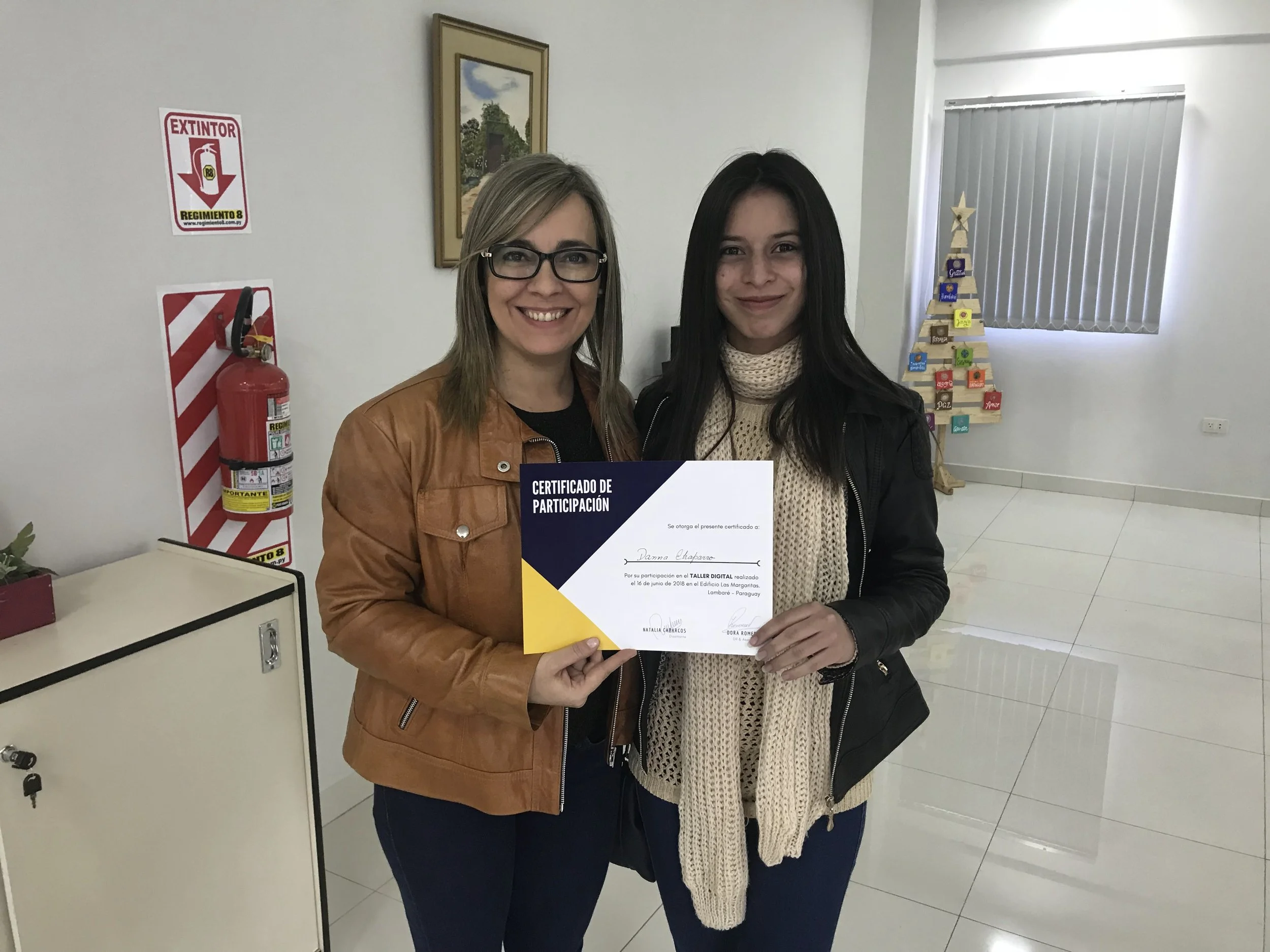Dos mujeres sonrientes sosteniendo un certificado de participación en un espacio interior con decoración navideña y un extintor en la pared.
