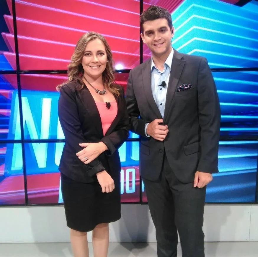 Una mujer y un hombre posando juntos en un estudio de noticias, ambos con ropa formal y sonriendo.