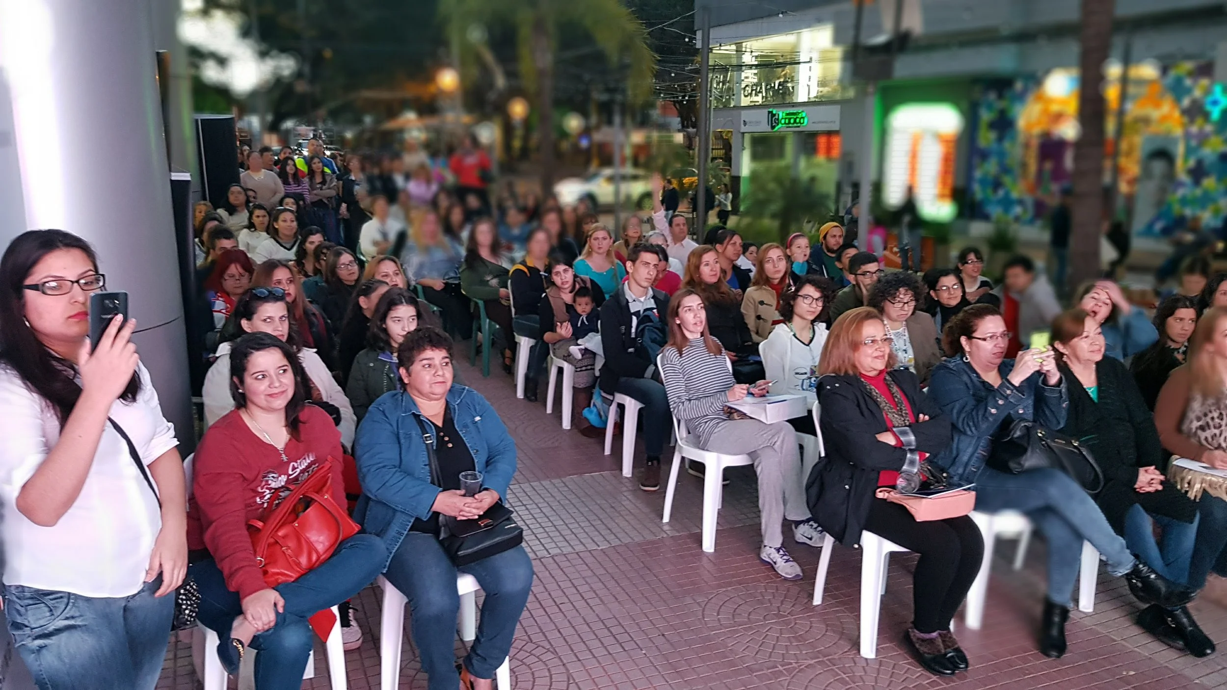 Grupo de personas asistiendo a un evento público al aire libre en una zona peatonal, algunas tomando fotos o videos con sus teléfonos, en un ambiente urbano y festivo.