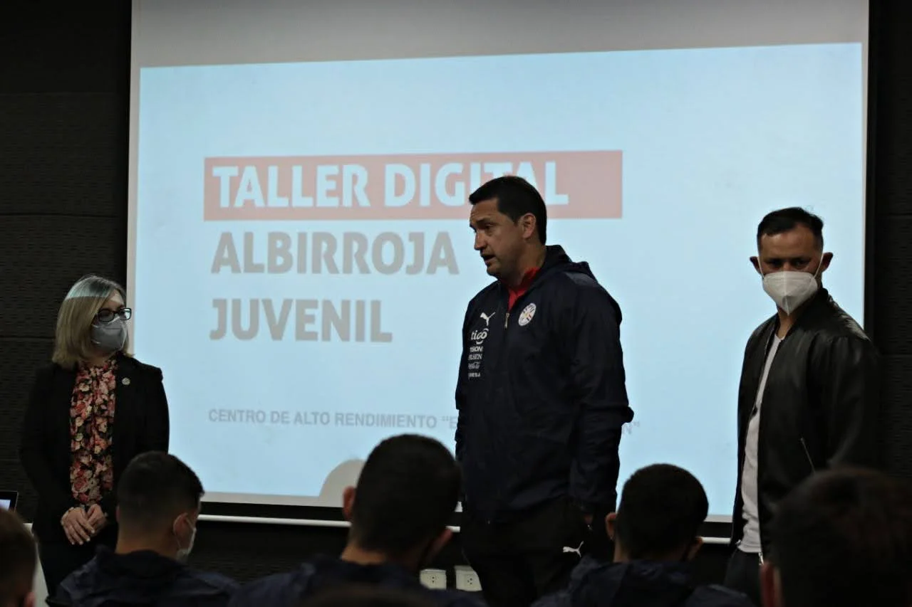 Una sala con tres personas de pie frente a un proyector. En la pantalla se lee 'Taller Digital Albirroja Juvenil'.