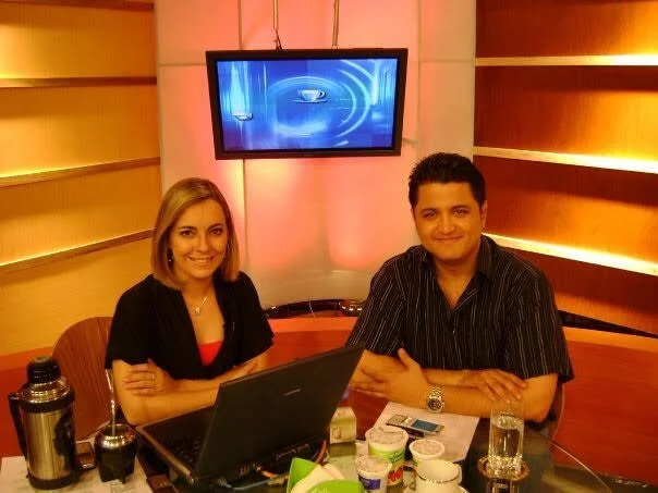 Dos personas, un hombre y una mujer, sentados en una mesa de estudio con una laptop, en un estudio de televisión.