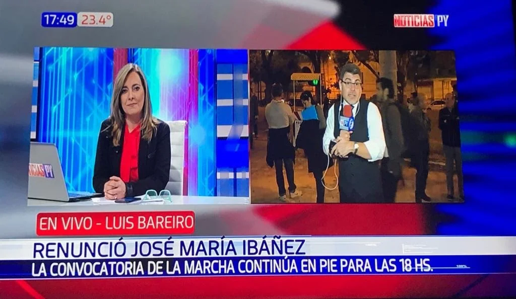 Pantalla de televisión con un segmento de noticias en vivo que muestra a una reportera en un estudio y a un reportero en la calle, con un cartel en pantalla que indica "EN VIVO - LUIS BAREIRO" y otra línea que informa sobre la renuncia de José María