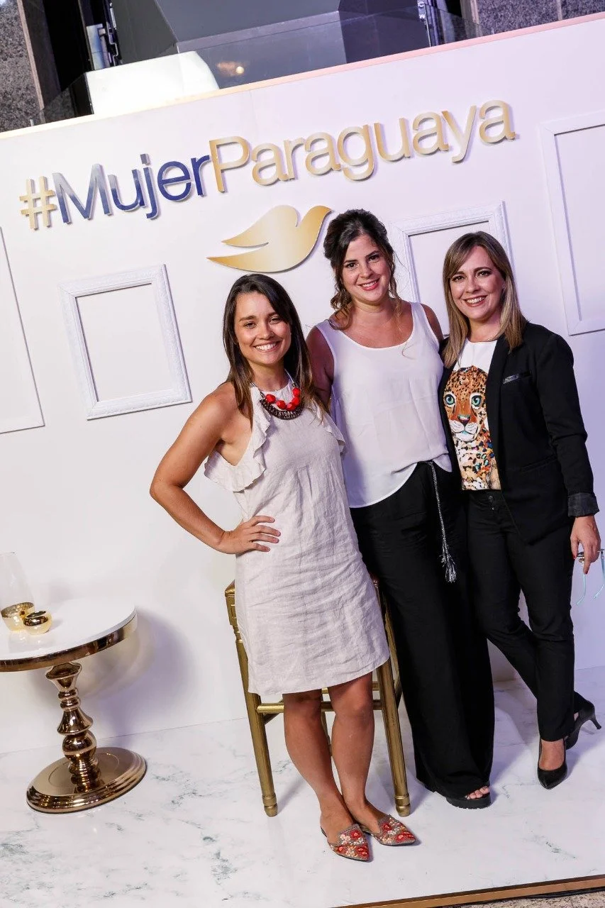 Tres mujeres sonriendo frente a un fondo blanco con un hashtag #MujerParaguaya y un símbolo de ave dorada, en un evento.