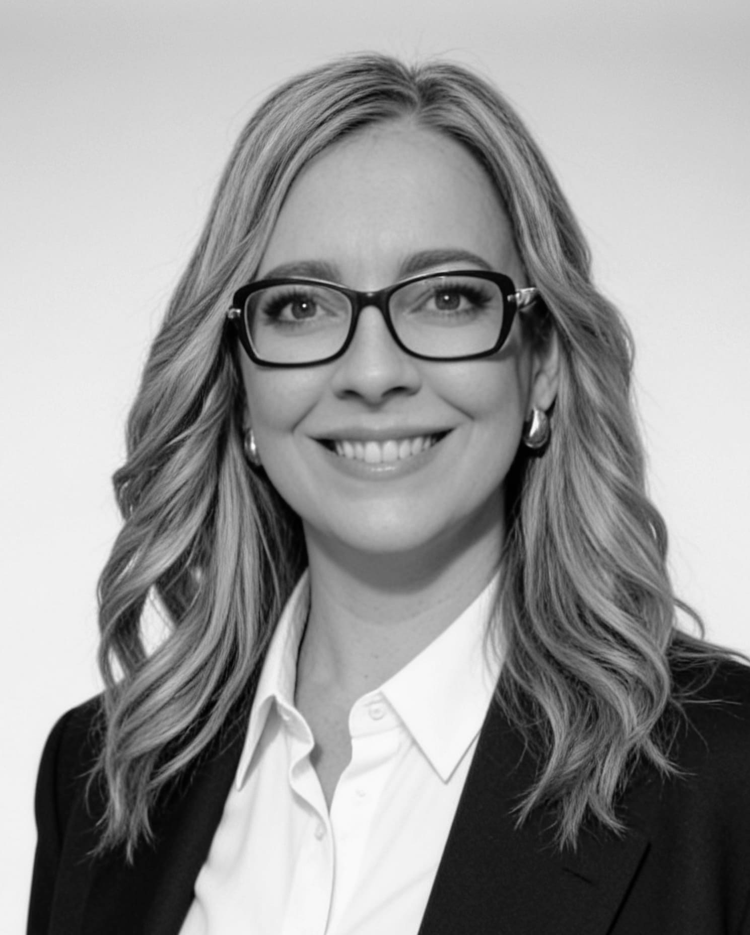 Retrato en blanco y negro de una mujer con gafas, cabello largo y rizado, sonriendo y vestida con saco y camisa blanca.