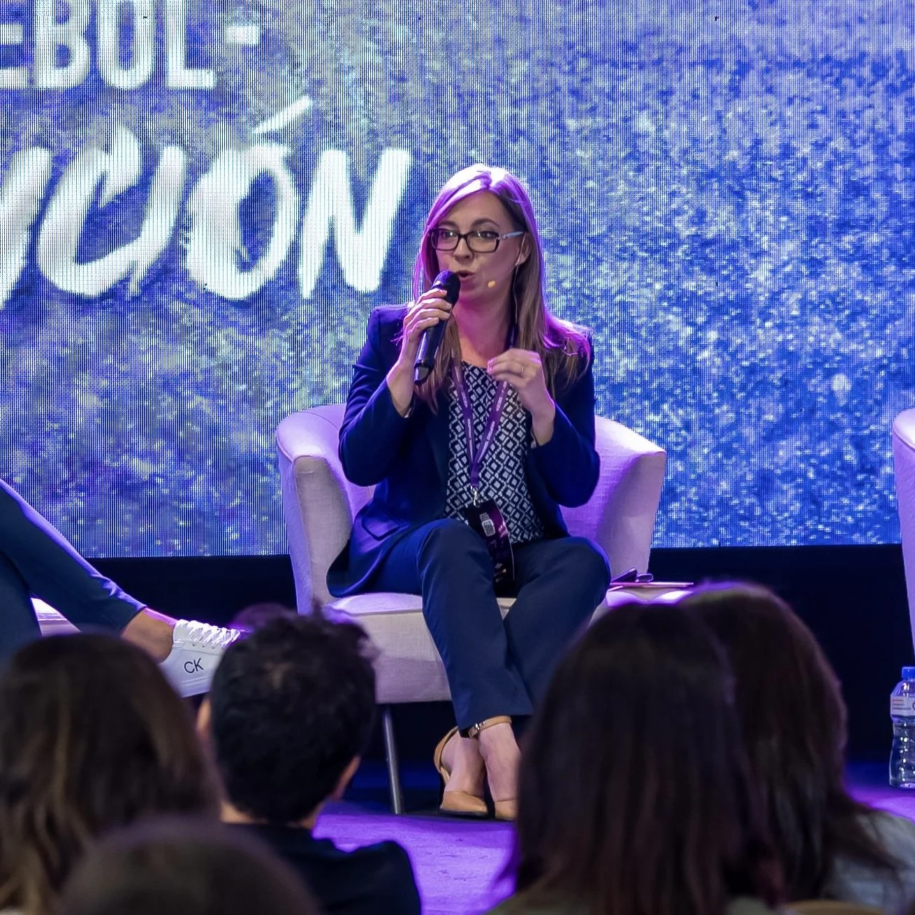 Una mujer sentada en una silla, hablando con un micrófono en un evento o conferencia, con un fondo brillante y letras visibles.