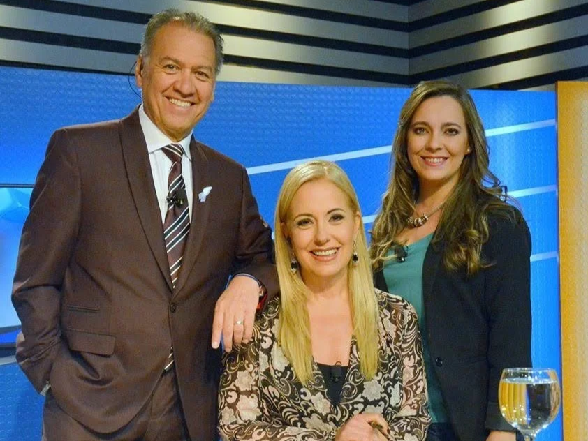 Tres presentadores en un programa de televisión, dos mujeres y un hombre, en un estudio con fondo azul.