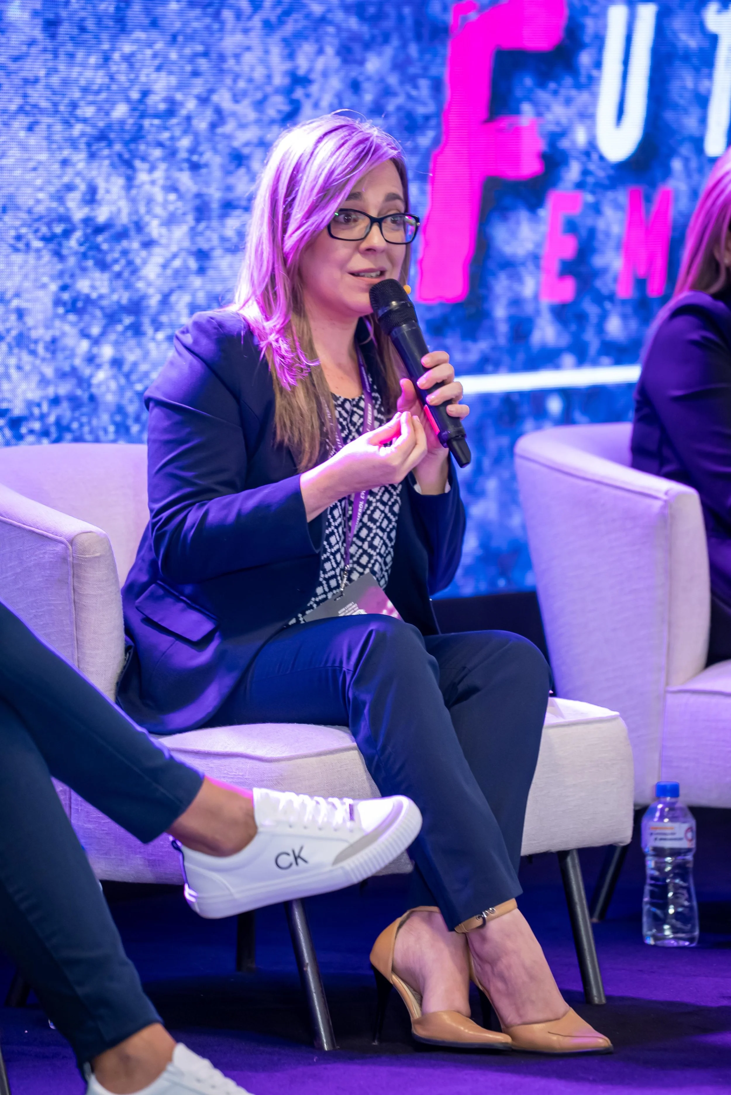 Mujer con gafas y cabello rubio, vestida con traje azul y tacones beige, hablando en un micrófono en una conferencia. Al fondo, un cartel con letras en rosa y azul.