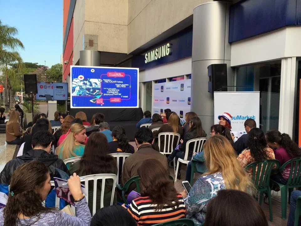 Reunión de personas en un evento al aire libre frente a una tienda Samsung, con pantalla grande y cartelera promocional.