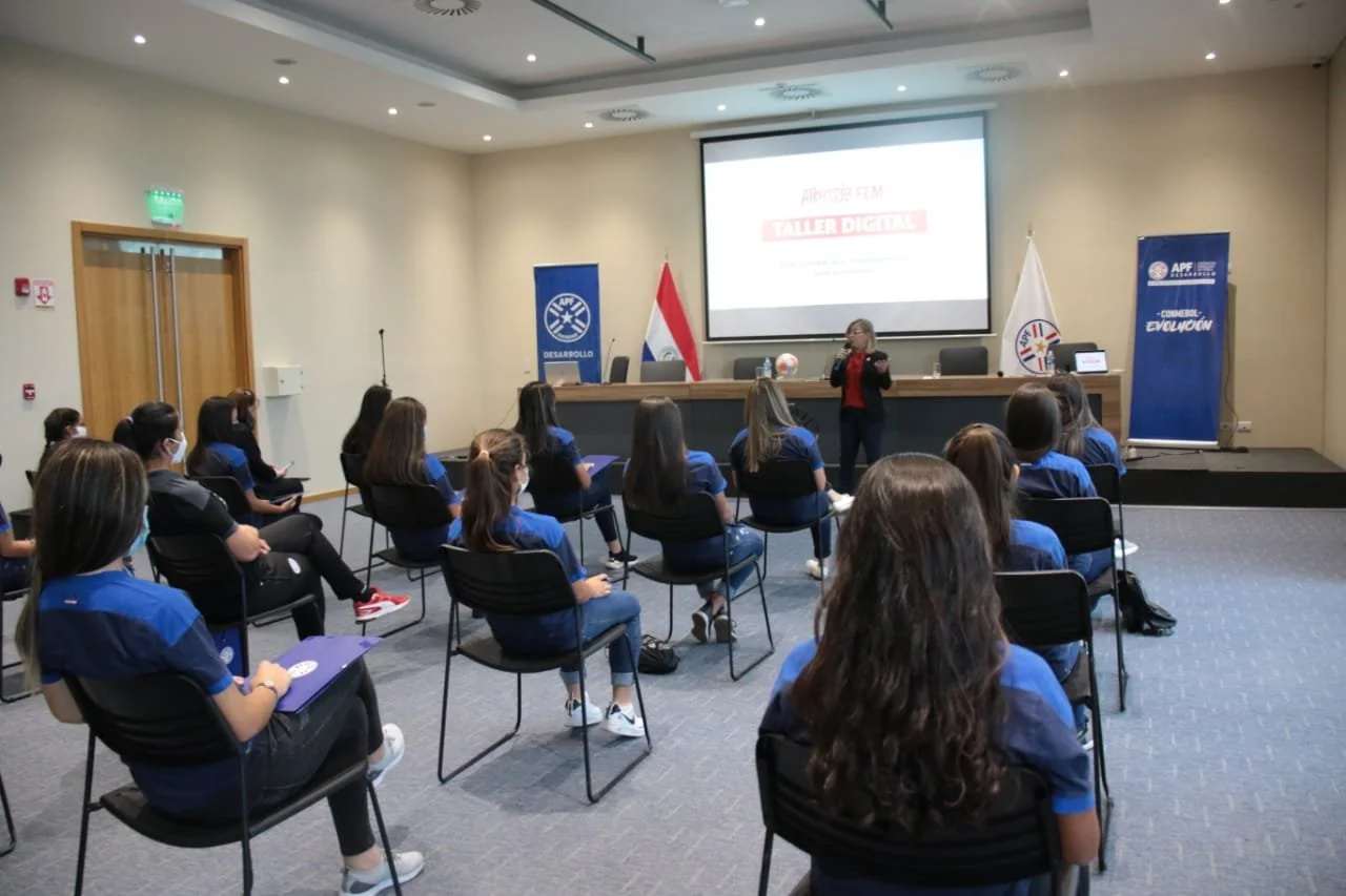 Una sala de conferencias con varias estudiantes sentadas usando mascarillas, atendiendo a una presentadora que está de pie frente a una pantalla grande. La pantalla muestra el título 'Albergue FEM Taller Digital' y hay banderas y banners al fondo.
