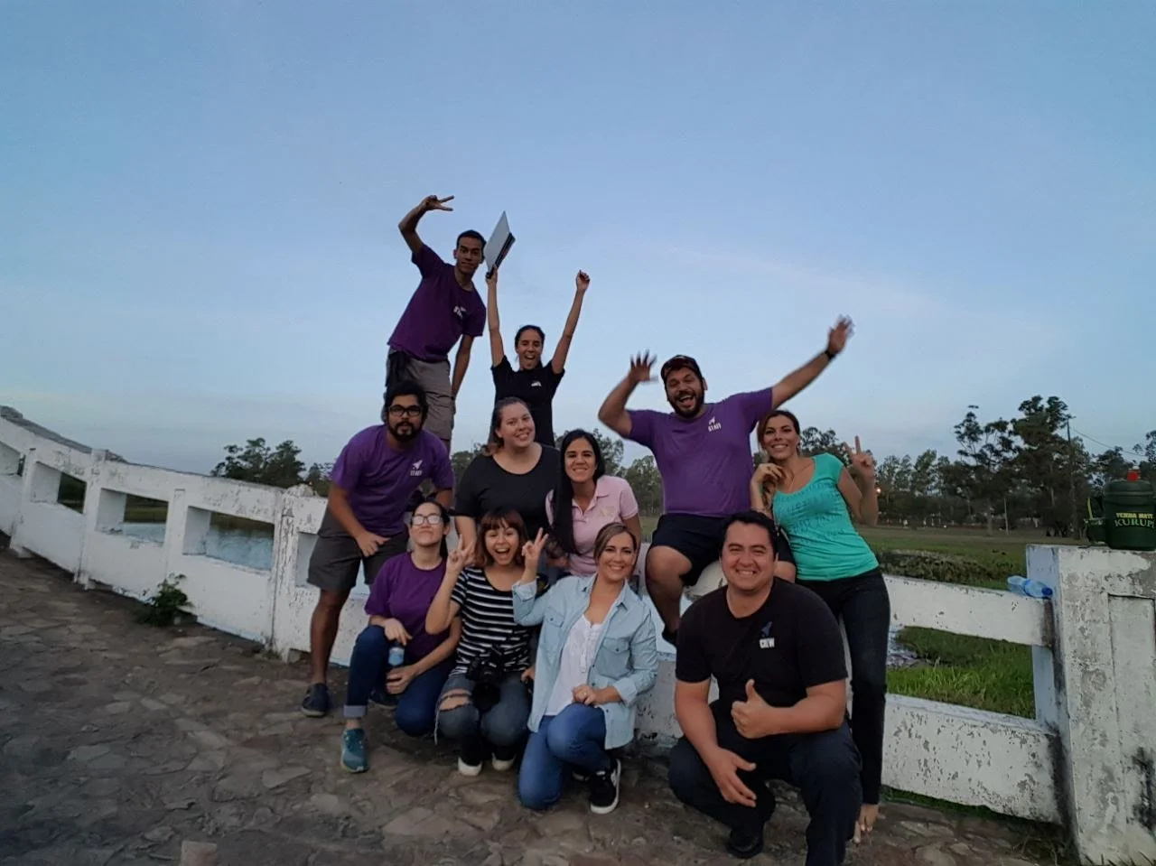 Grupo de personas sonriendo y posando al aire libre durante el atardecer, algunos haciendo gestos y otros levantando las manos, con fondo de campo y árboles.