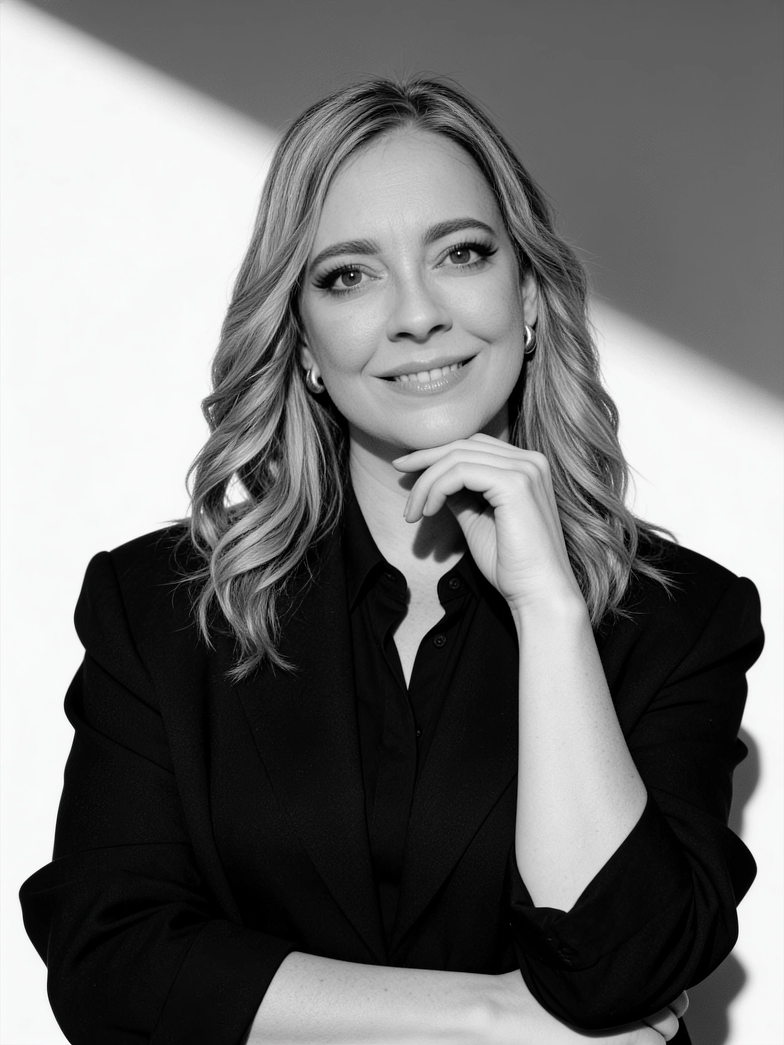 Retrato en blanco y negro de una mujer con cabello rubio rizado y largo, sonriendo levemente, con la mano bajo la barbilla, usando una chaqueta negra y una blusa.