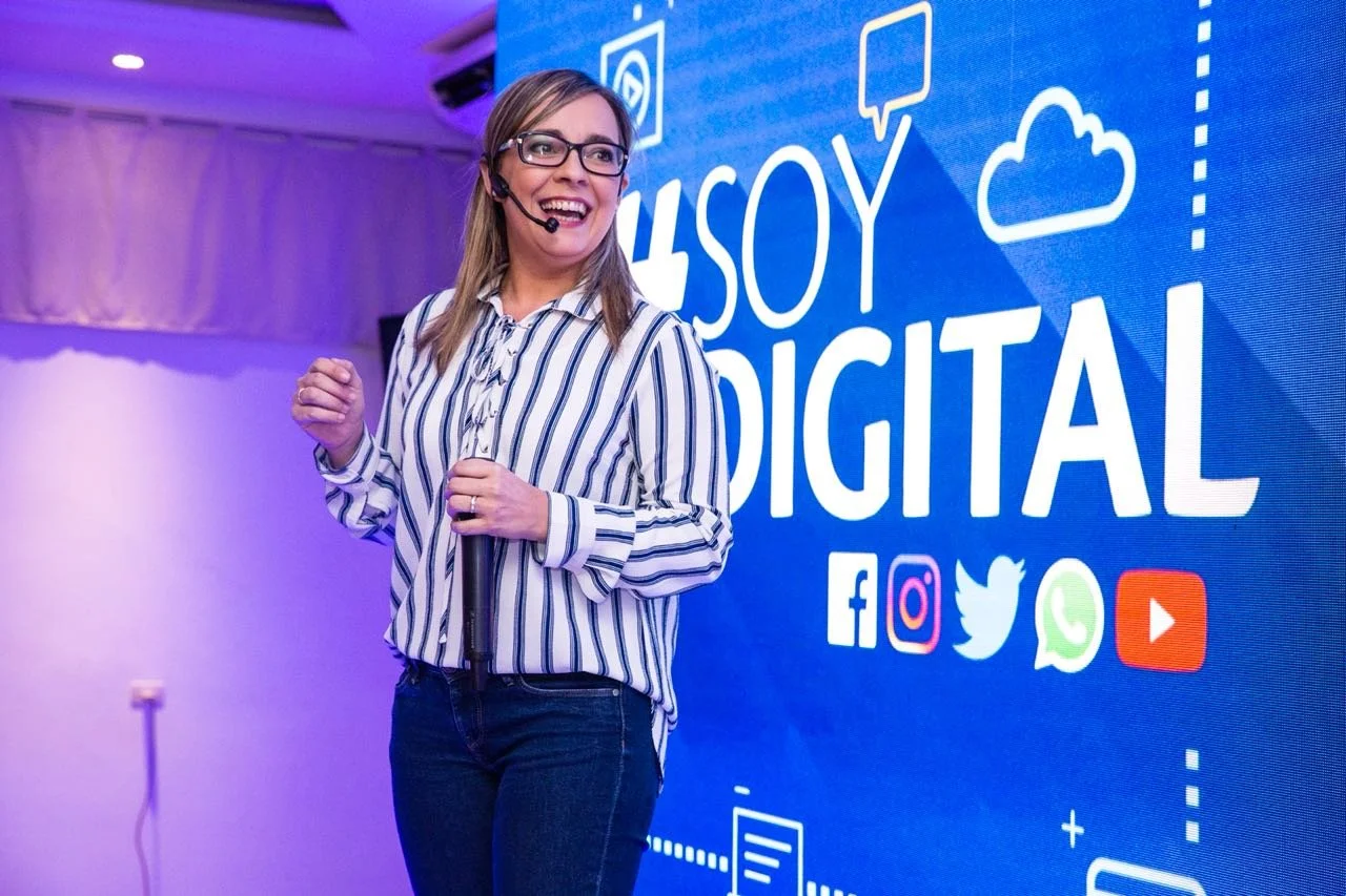 Mujer en una conferencia sobre tecnología, con una sonrisa, usando un auricular y sosteniendo un micrófono, frente a una pantalla que dice '¡Yo soy digital' con iconos de redes sociales.