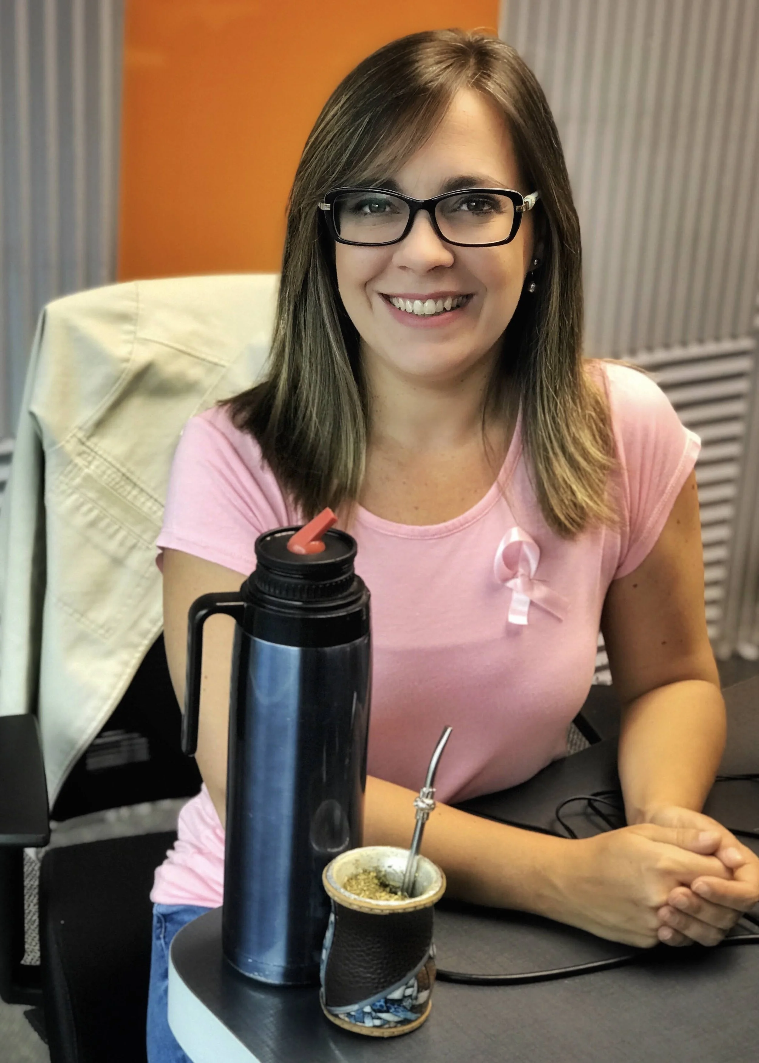 Mujer sonriendo, lleva gafas y camiseta rosa, sentada en oficina con una termos y mate en la mesa.