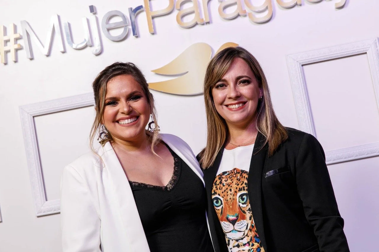 Dos mujeres sonriendo, de cabello castaño, en un evento con fondo blanco y texto 'Mujer Paraguay' en letras grandes.