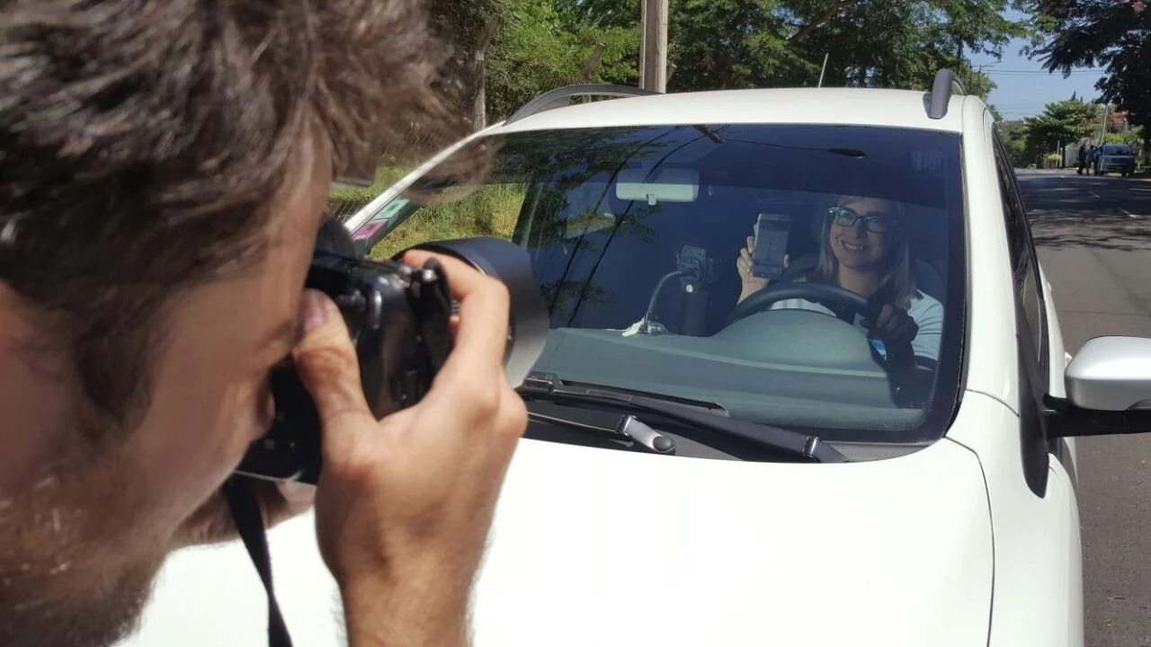 Una persona al frente toma una fotografía a otra que está conduciendo un automóvil blanco y le sonríe desde el interior del vehículo, en una calle con árboles y otros autos al fondo.