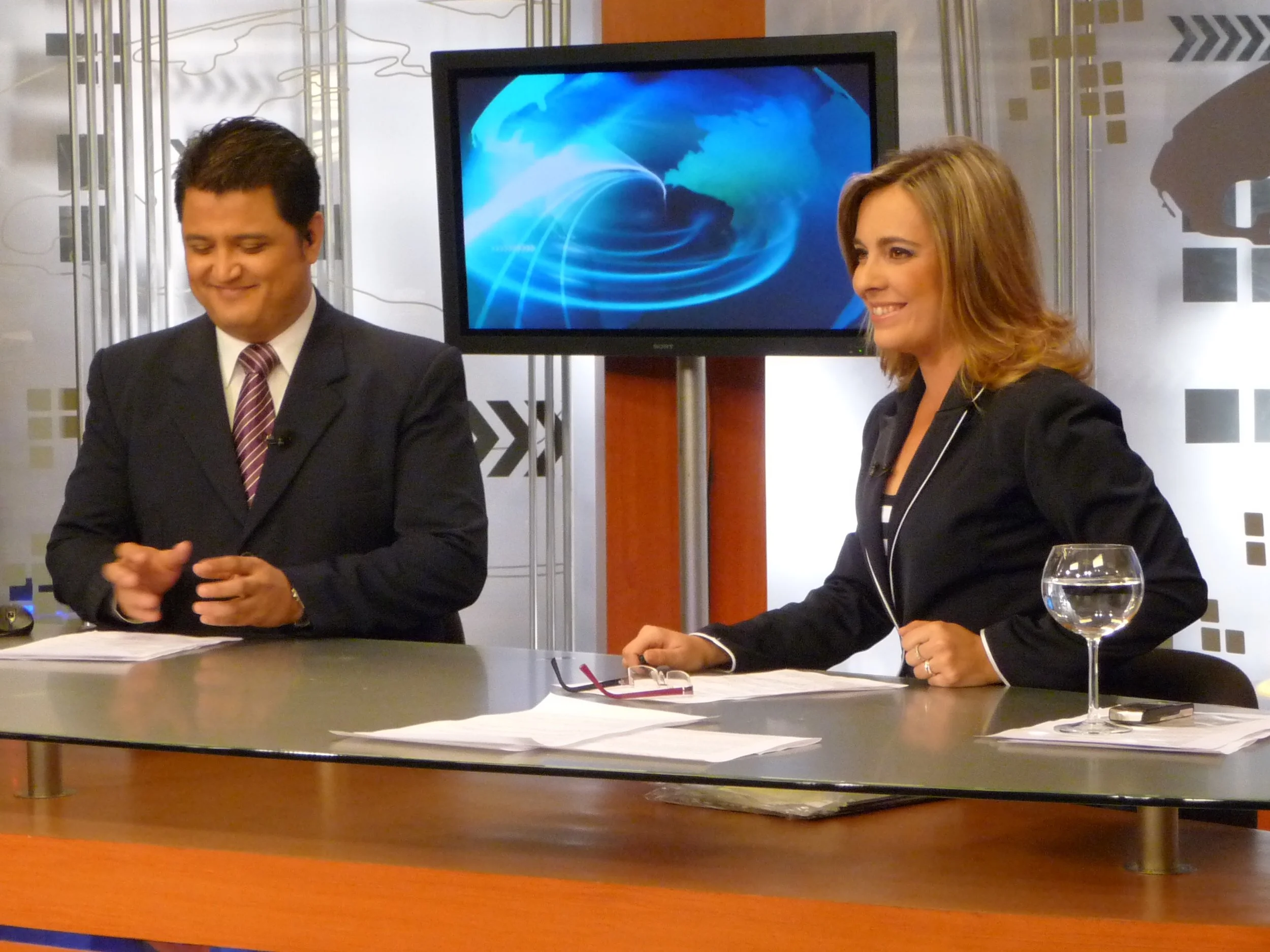 Dos presentadores, un hombre y una mujer, en un estudio de televisión, sentado tras una mesa con documentos y un vaso con agua.