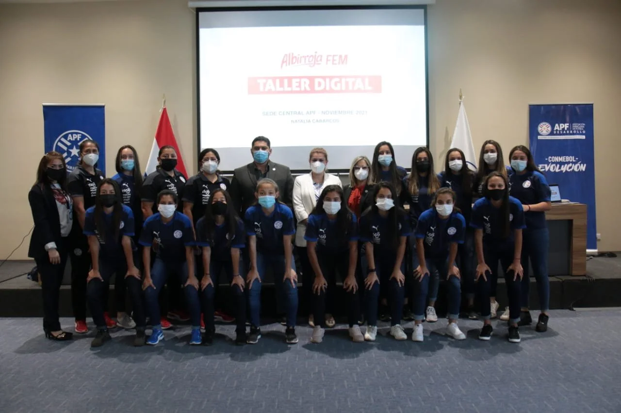 Grupo de personas en una conferencia, con un proyector que muestra un slide con texto en español. Algunos llevan camisetas y mascarillas, y están en un escenario con banderas y un fondo azul con logotipos de APF y una pantalla que dice 'Taller Digita