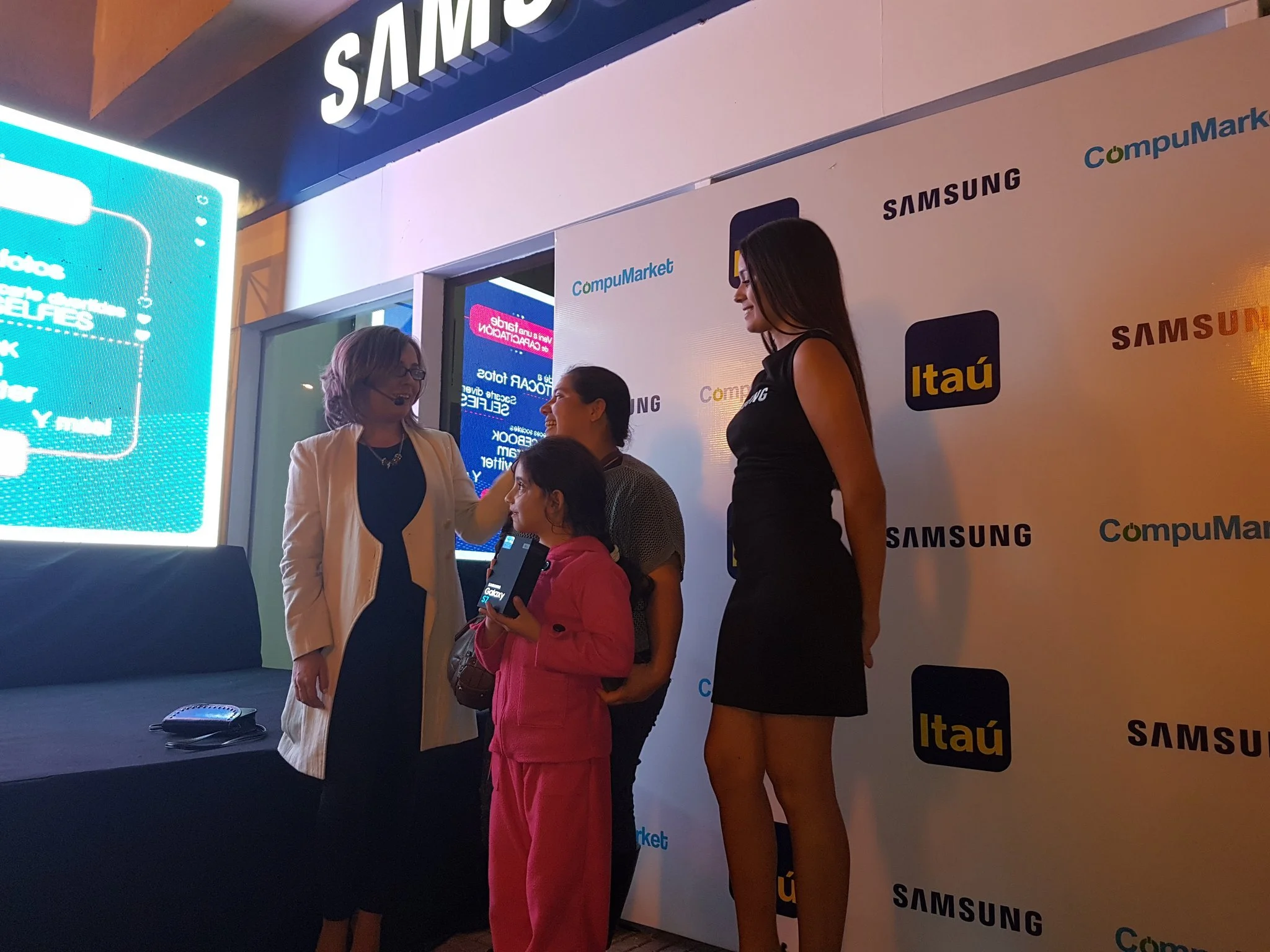 Una mujer presenta un teléfono móvil a una niña en un evento de tecnología, con un fondo que muestra logotipos de Samsung, Itaú y CompuMarket.