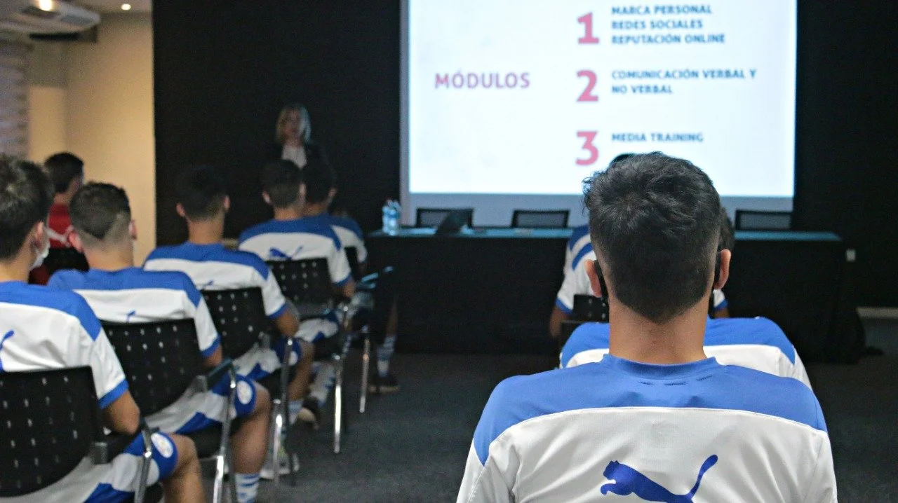 Estudiantes en uniforme azul y blanco en una sala de clases, viendo una presentación en una pantalla grande que muestra módulos de un curso o entrenamiento.