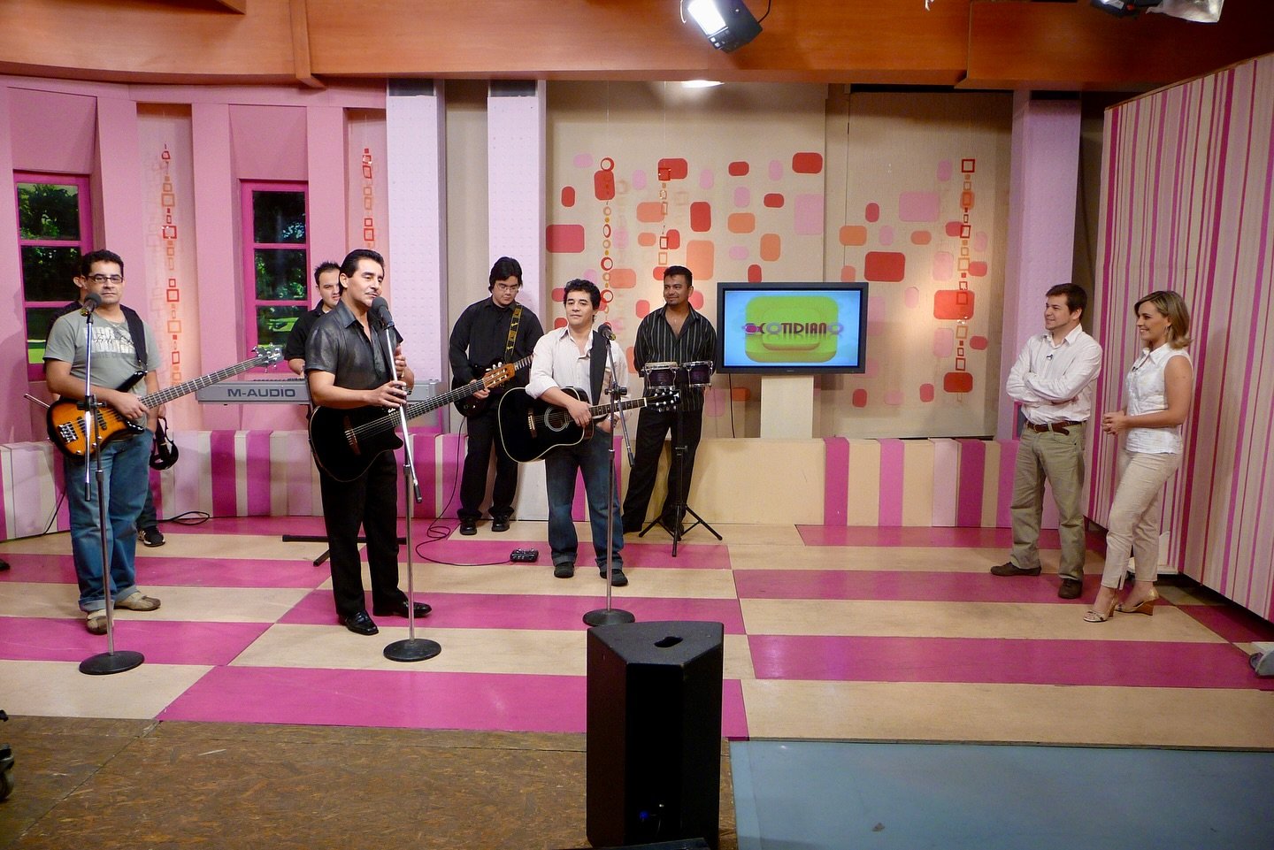 Un grupo de músicos tocando en un set de televisión con una presentadora y un presentador en un estudio de grabación colorido.