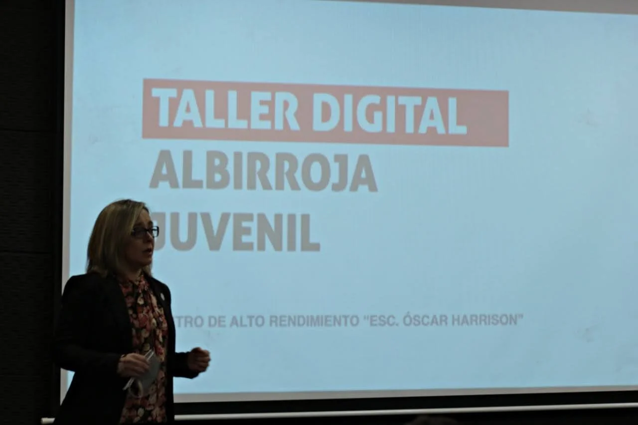 Presentadora en una conferencia con una diapositiva que dice "Taller Digital Albirroja Juvenil" y que menciona un centro de alto rendimiento "Esc. Oscar Harrison".