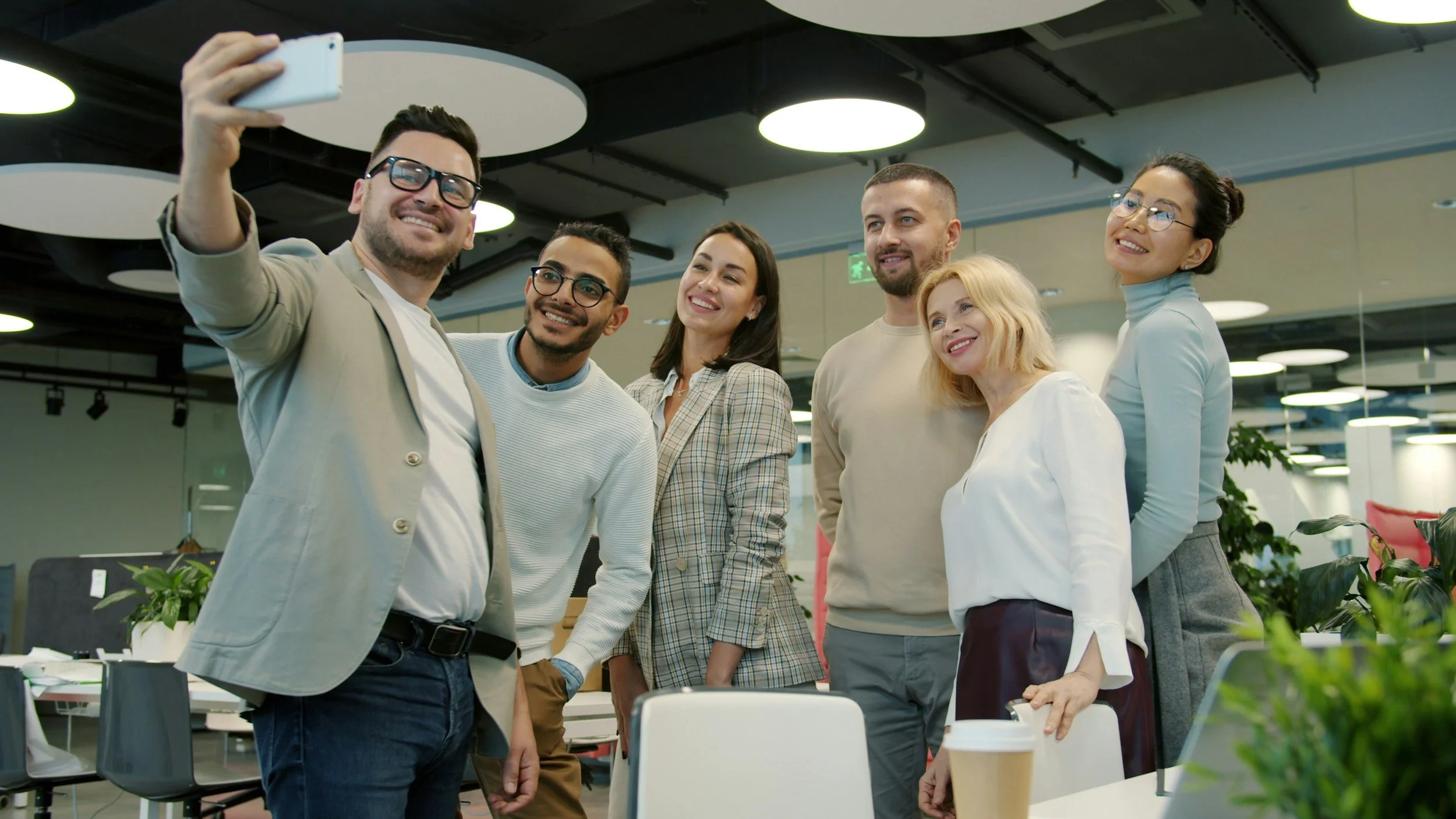 Group of six diverse young professionals taking a selfie in a modern office workspace, smiling and standing close together.