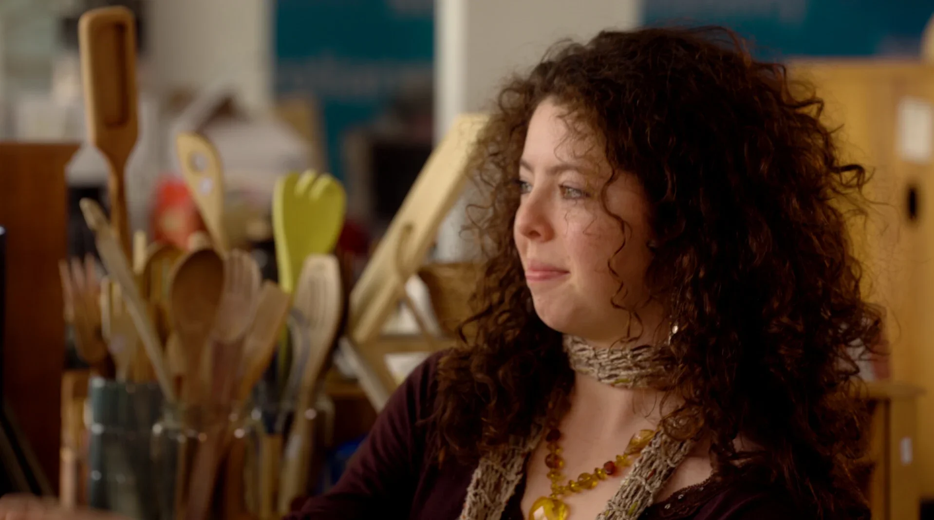 A woman with curly brown hair and light skin sitting at a counter in a kitchen, surrounded by utensils and kitchenware.