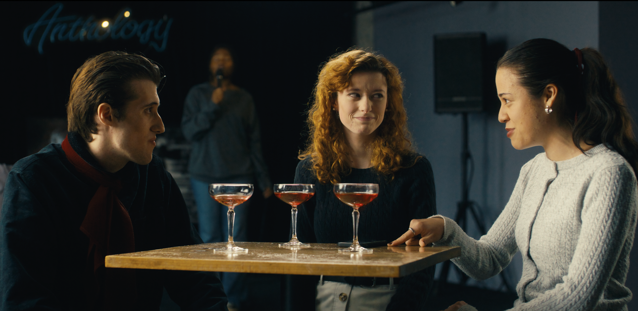 A scene from Lemon Time's short dotdotdot" Three young people—two women and one man—are sitting at a table with three cocktails in a dimly lit bar or restaurant.