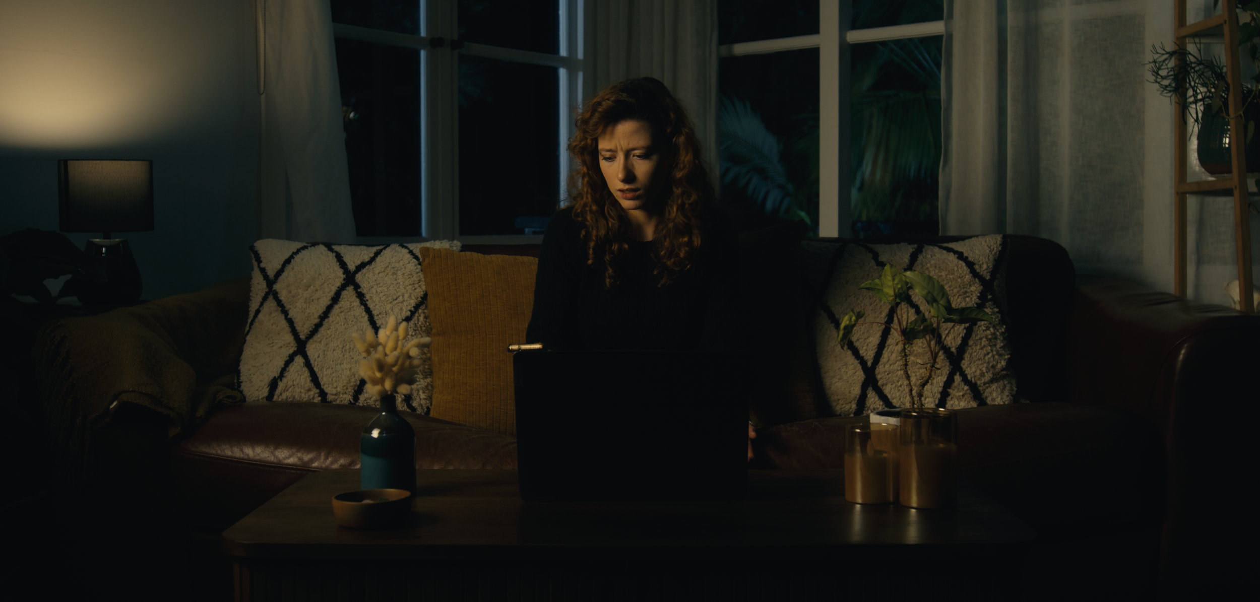 A scene from Lemon Time's short "dotdotdot" where a woman with curly hair is sitting on a couch at night, working on a laptop. The cozy living room has a lamp, plants, and decorative pillows, with a window showing the dark outside.
