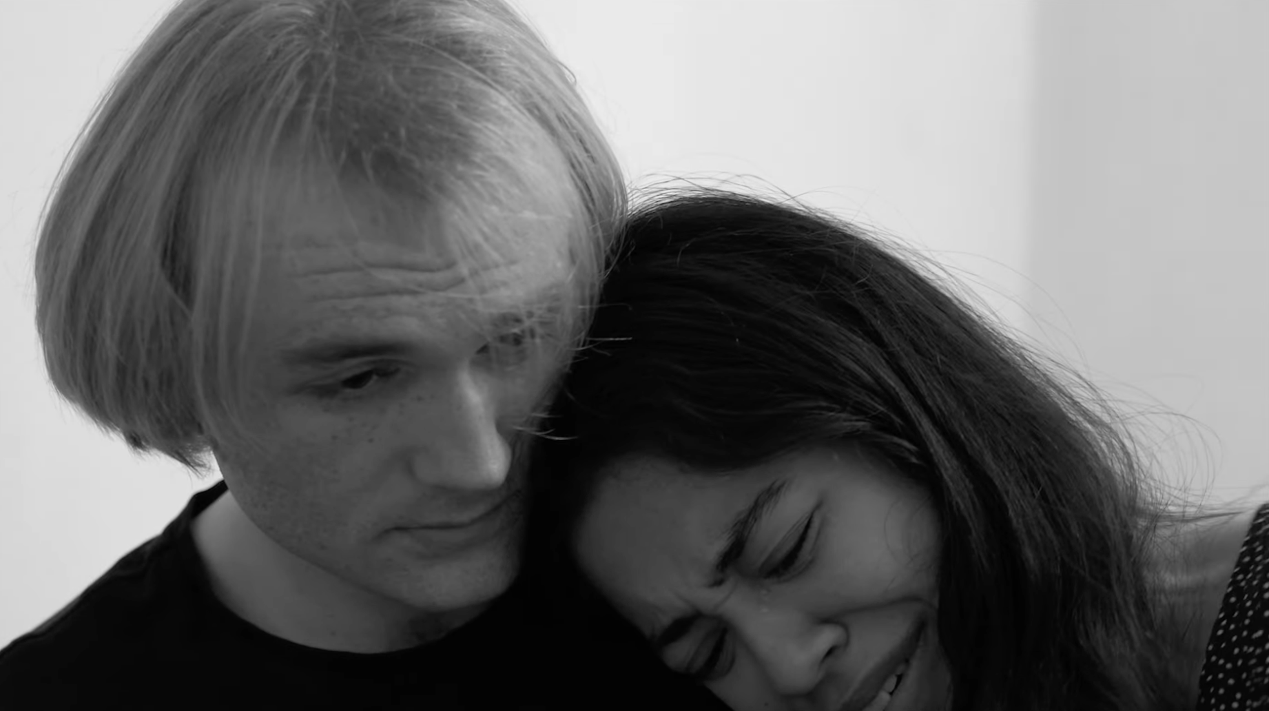 A black and white close-up photograph of two women embracing. The woman on the left has light skin, freckles, and short light-colored hair, while the woman on the right has darker skin and long dark hair. They are resting their heads against each other, with the woman on the right smiling softly.