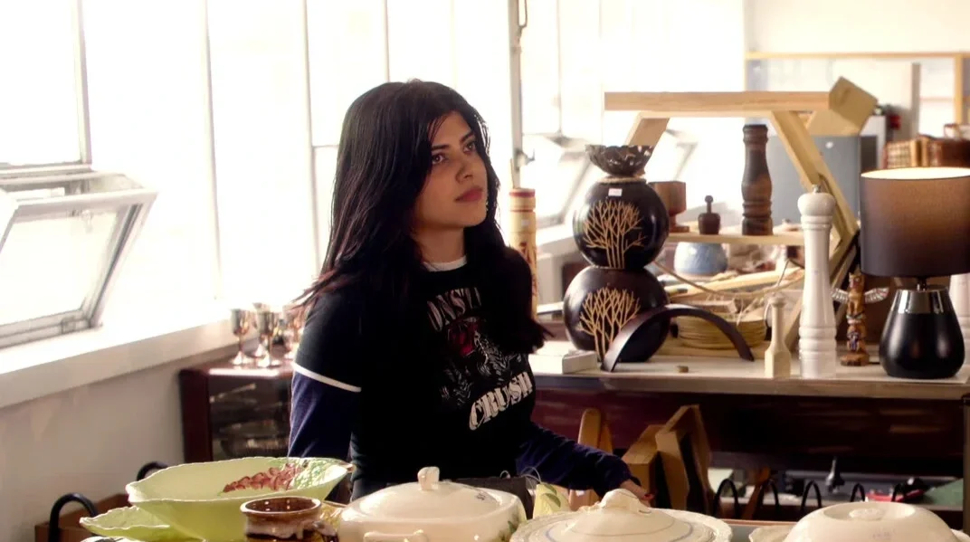 A woman with long dark hair wearing a black and white shirt sitting at a cluttered table in a bright room with large windows.