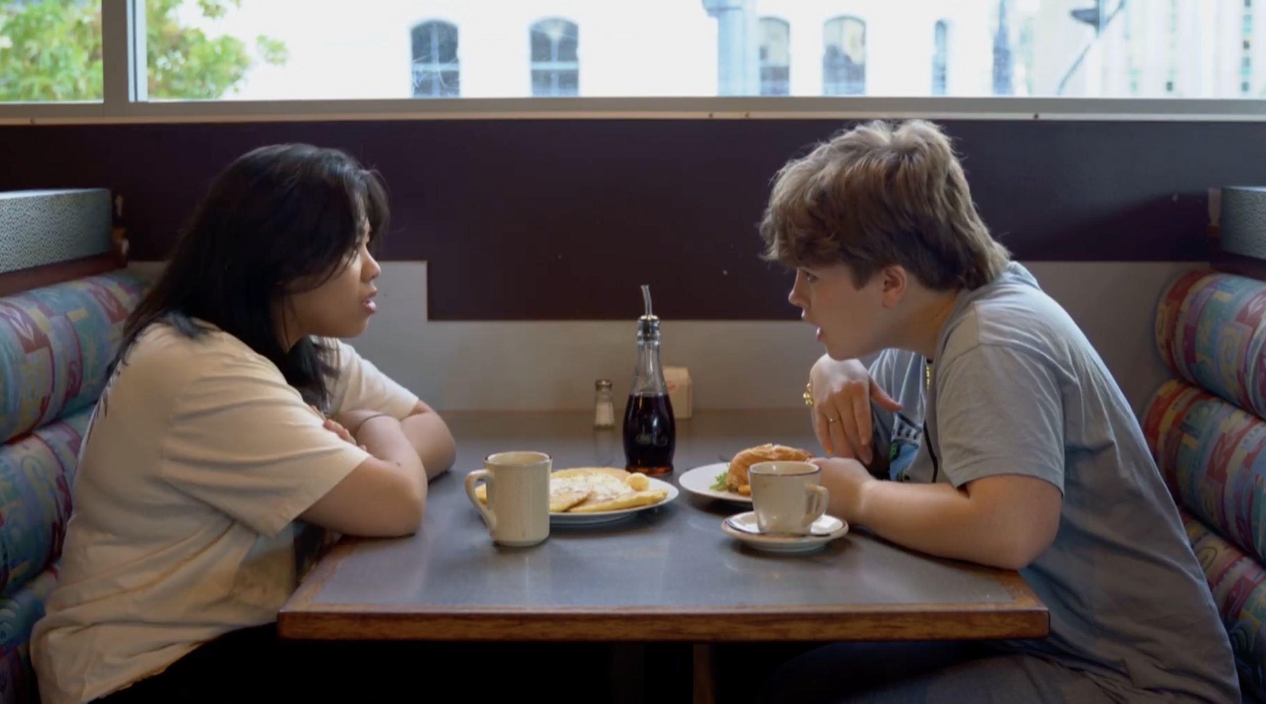 Two people, a woman and a man, are sitting across from each other in a booth at a restaurant, having a conversation during breakfast. The woman has black hair and wears a light-colored shirt, the man has light brown, tousled hair and wears a gray T-shirt. On the table are plates of breakfast food, including eggs and fried items, cups of coffee, and a glass bottle of soda.