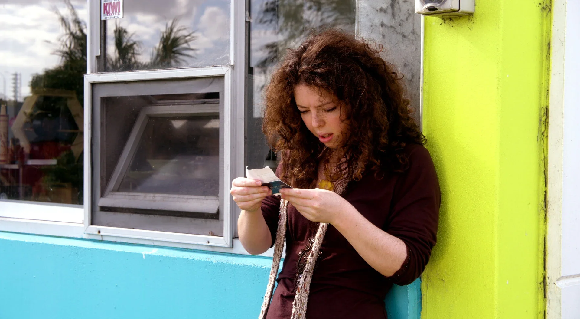 A woman with curly brown hair, wearing a dark jacket, stands outside a colorful building, looking at a piece of paper or ticket in her hand. The building has a yellow, green, and blue exterior, with a window and a small white box mounted on the wall.