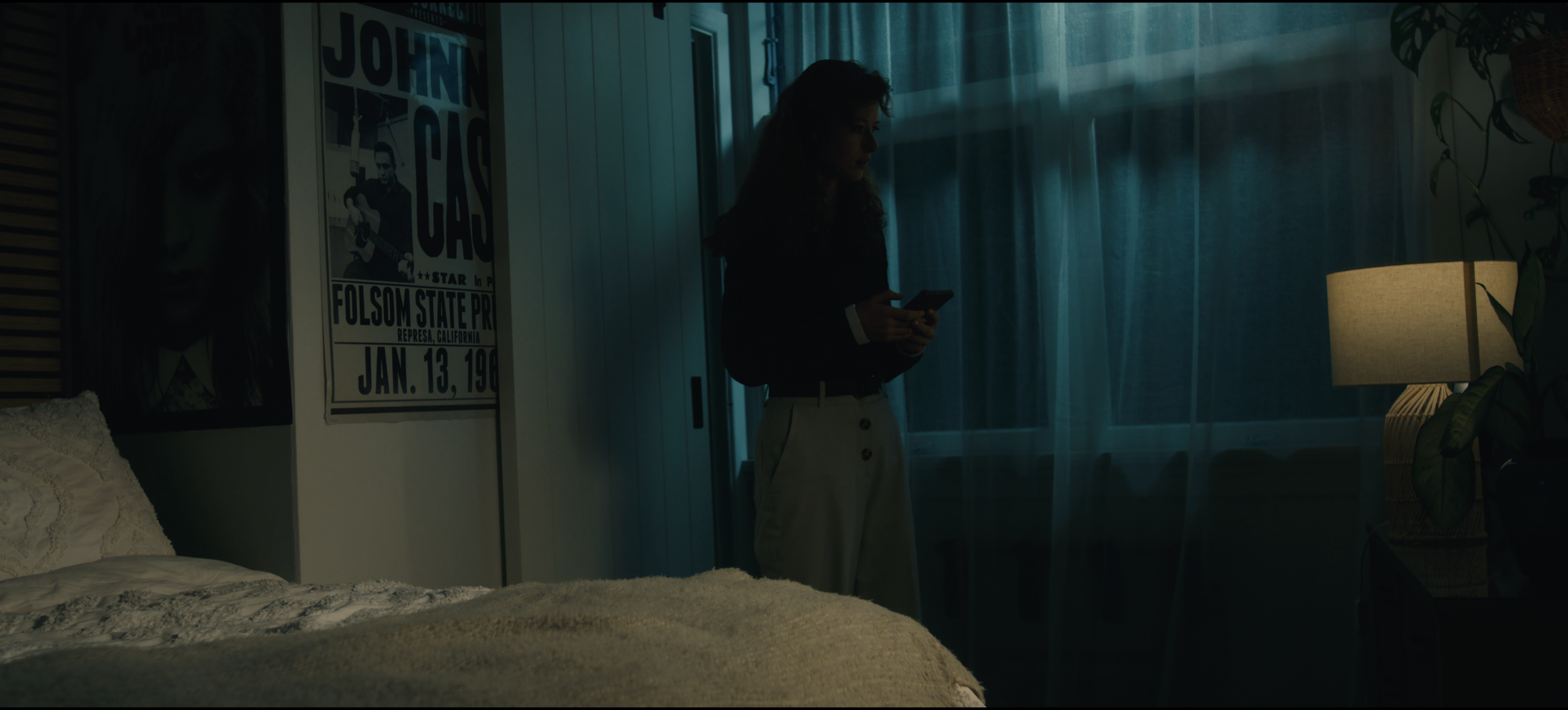A scene from Lemon Time's short "dotdotdot"  with a woman standing near a window, looking at her phone in a dimly lit room with posters on the wall, a lamp, and a plant.