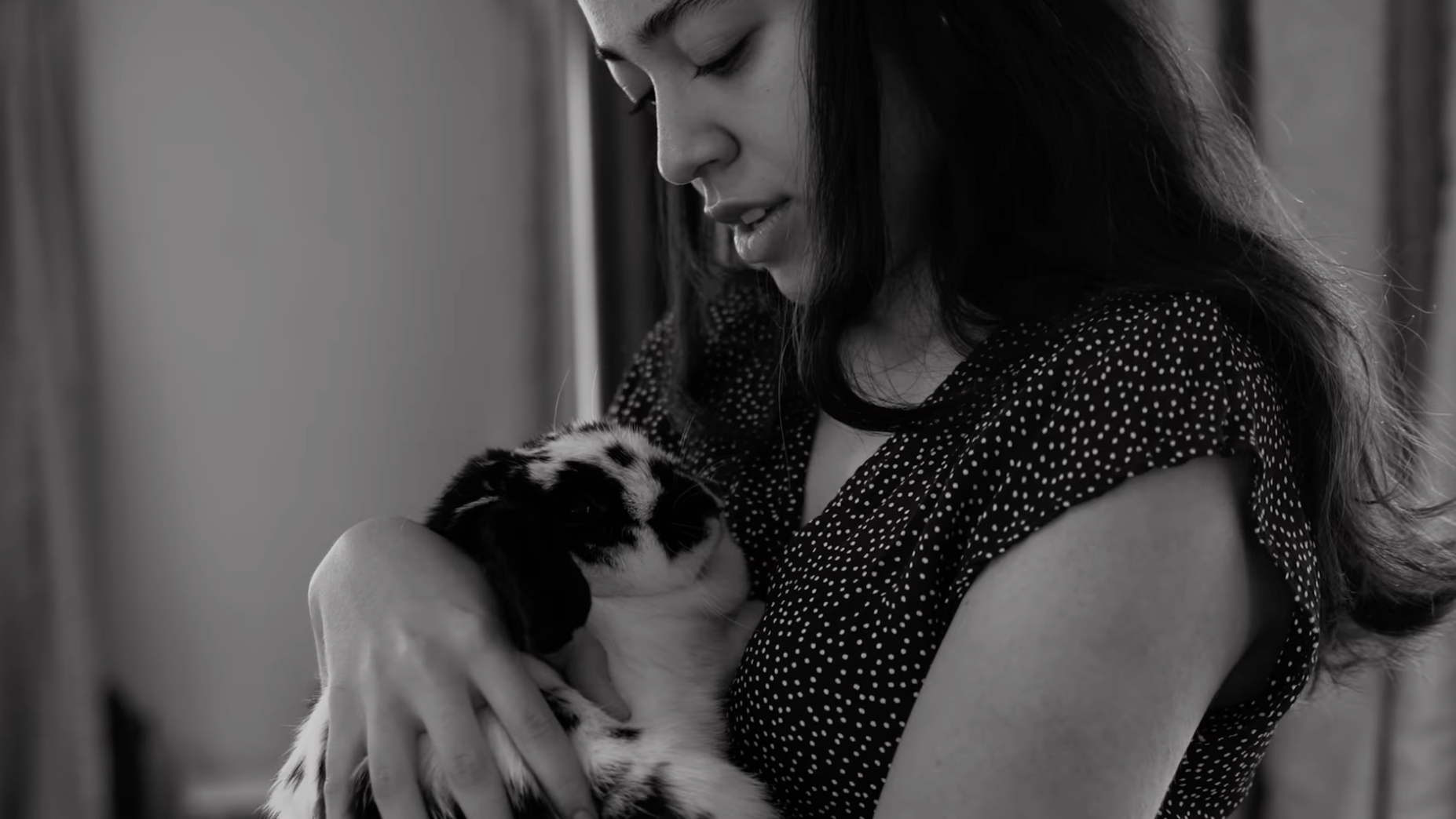 A woman with long dark hair, wearing a polka-dot shirt, holding and looking down at a small black and white kitten.