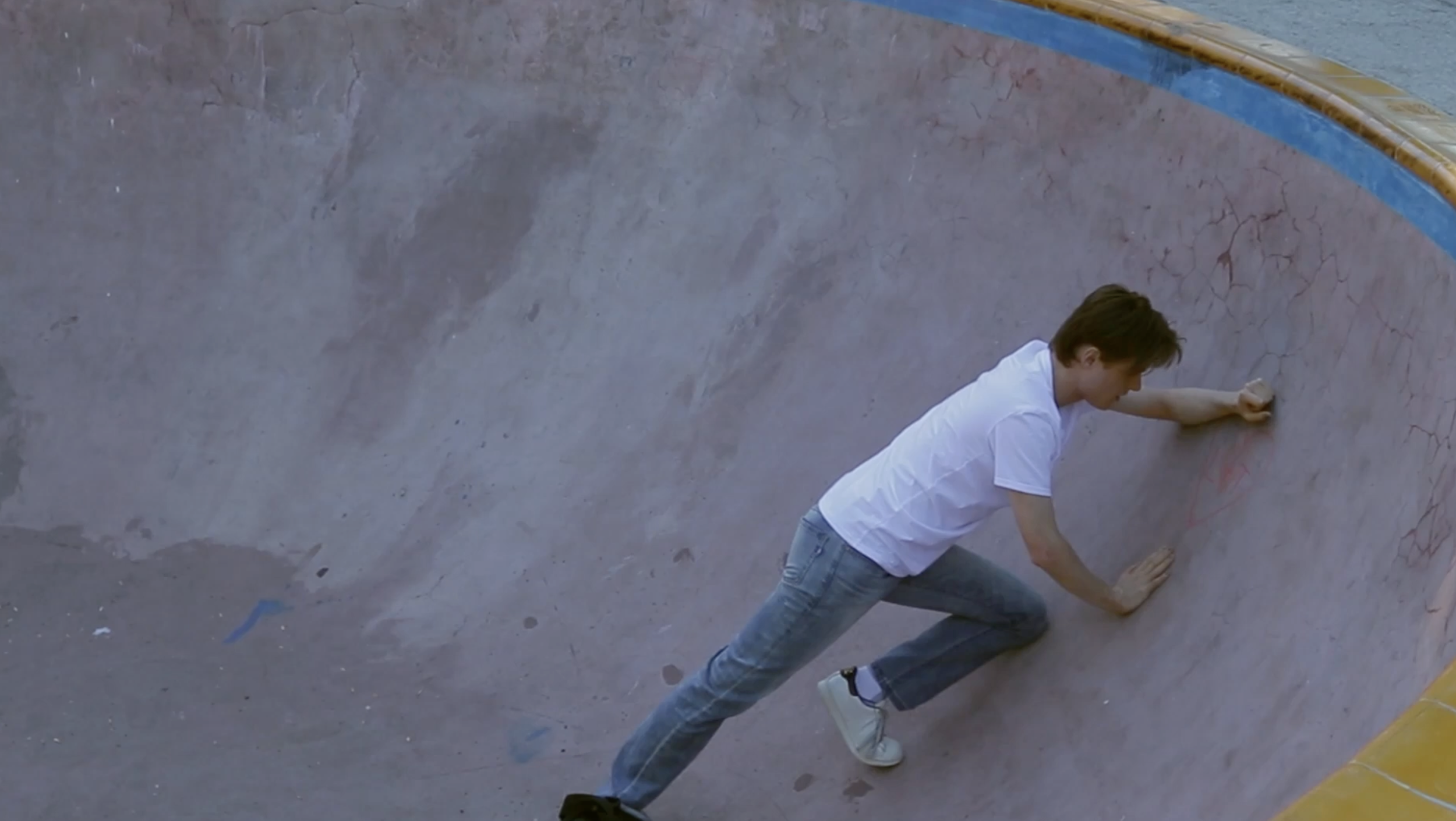 A person wearing a white shirt and jeans in a fractured concrete skateboard bowl, leaning on their hands.