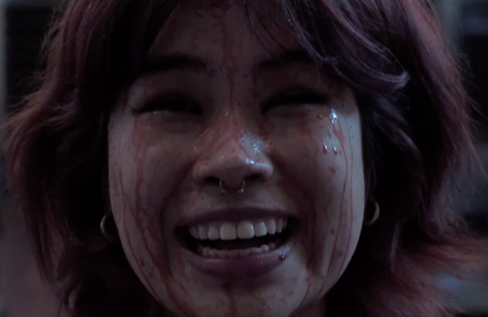 A close-up of a woman smiling with blood on her face, blood running down her forehead and cheeks, and a septum piercing, with dark hair and earrings.