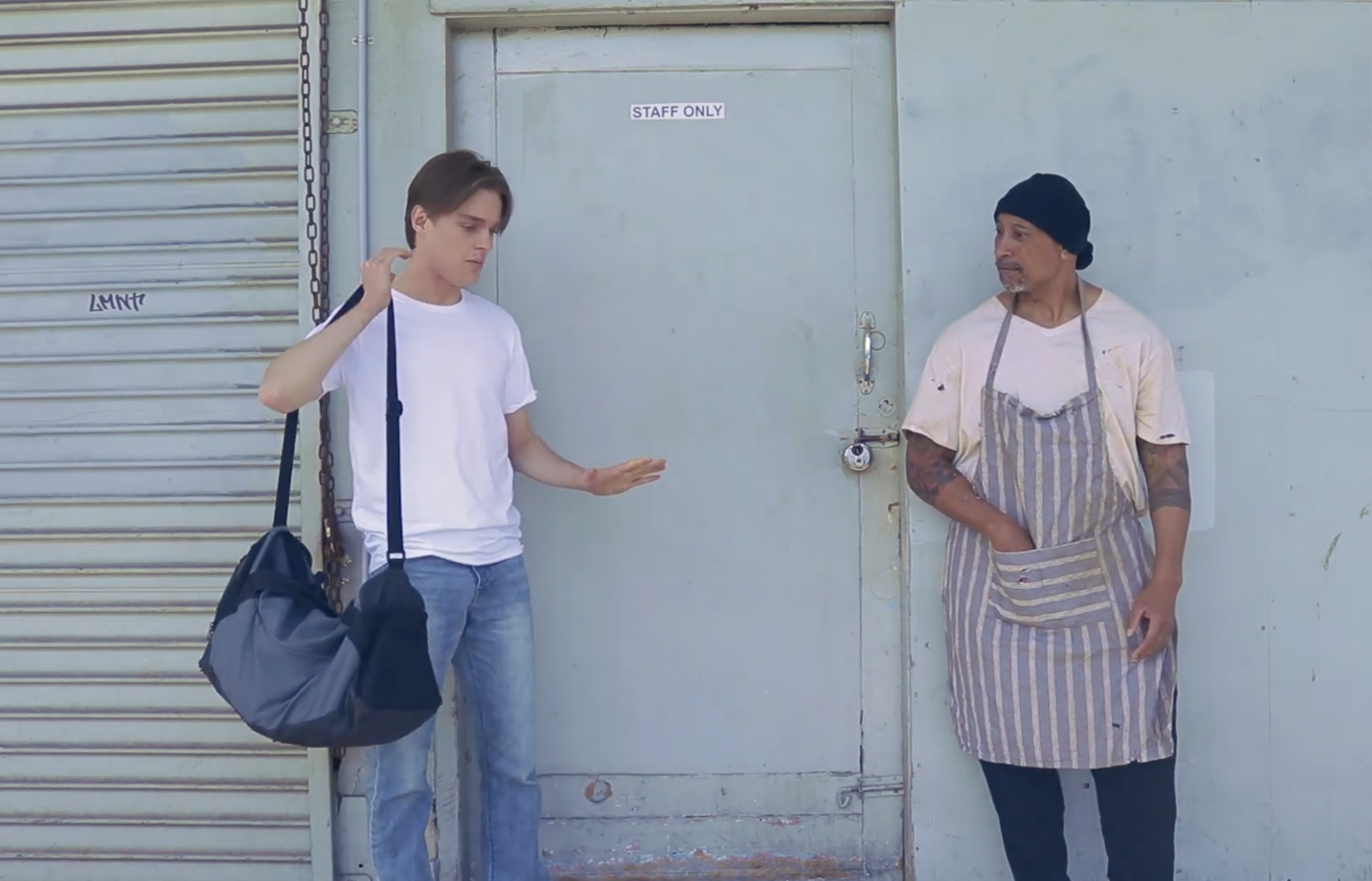 A young man with short brown hair, wearing a white t-shirt and carrying a black bag, is leaning against a closed metal shutter door with a 'STAFF ONLY' sign. He appears to be talking to an older man with a beard, wearing a black beanie, a white t-shirt with stains, and a striped apron, standing with his hands in his pockets.