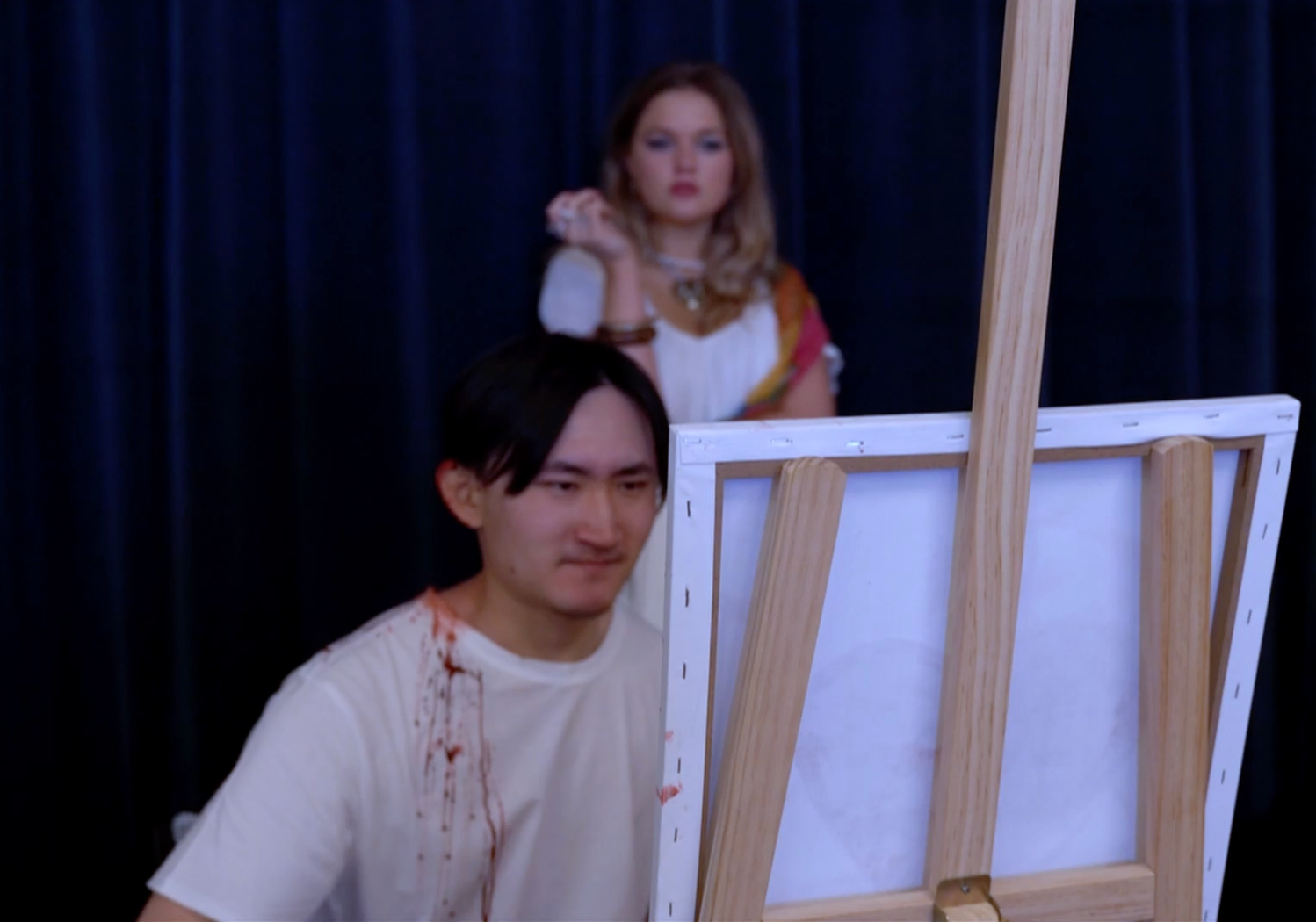 A man with fake bloodstains on his white shirt is sitting in front of a blank canvas on an easel, while a woman dressed in renaissance-style clothing stands in the background behind him, with a dark curtain as the backdrop.