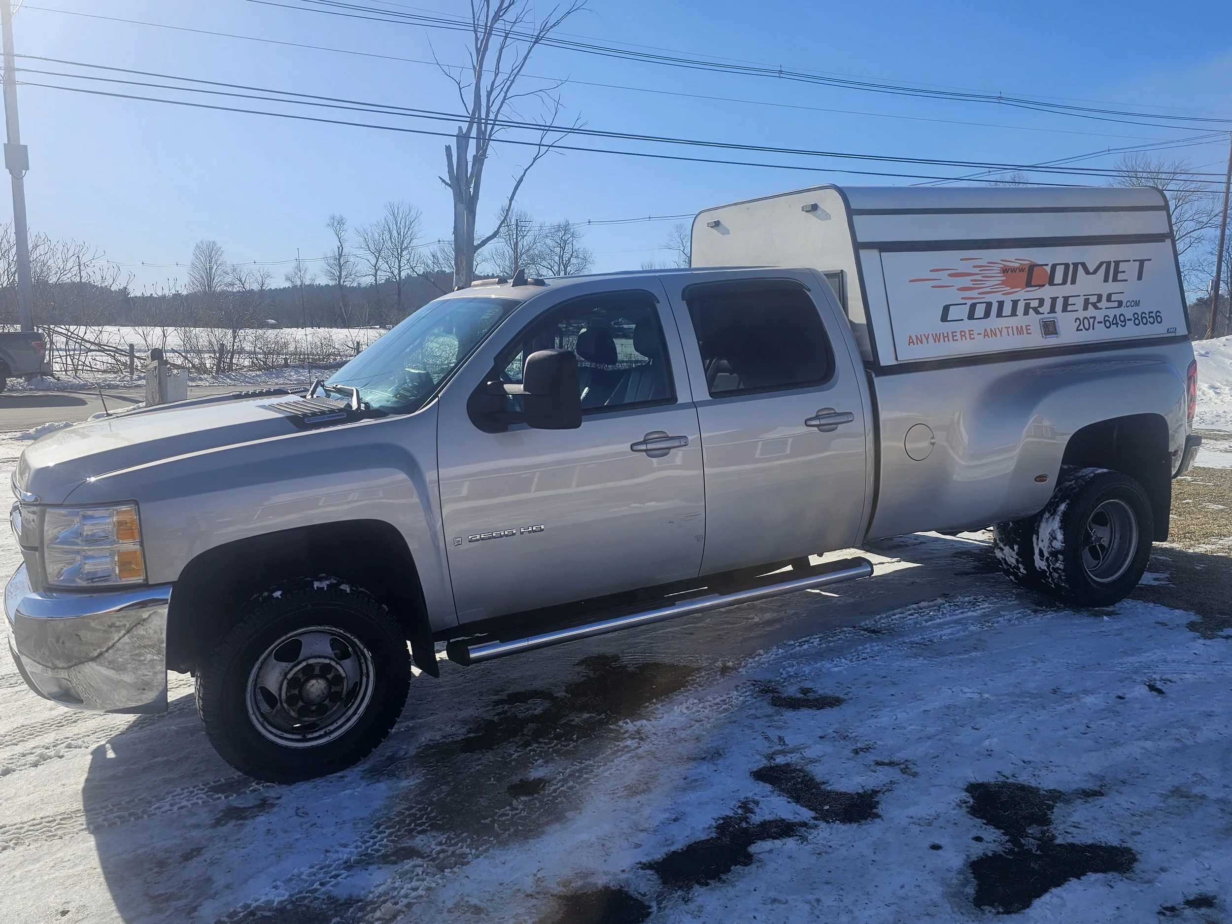 Silver pickup truck with a graphic box on the bed advertising Comet Couriers, parked on snow-covered ground under a blue sky, with leafless trees and power lines in the background.