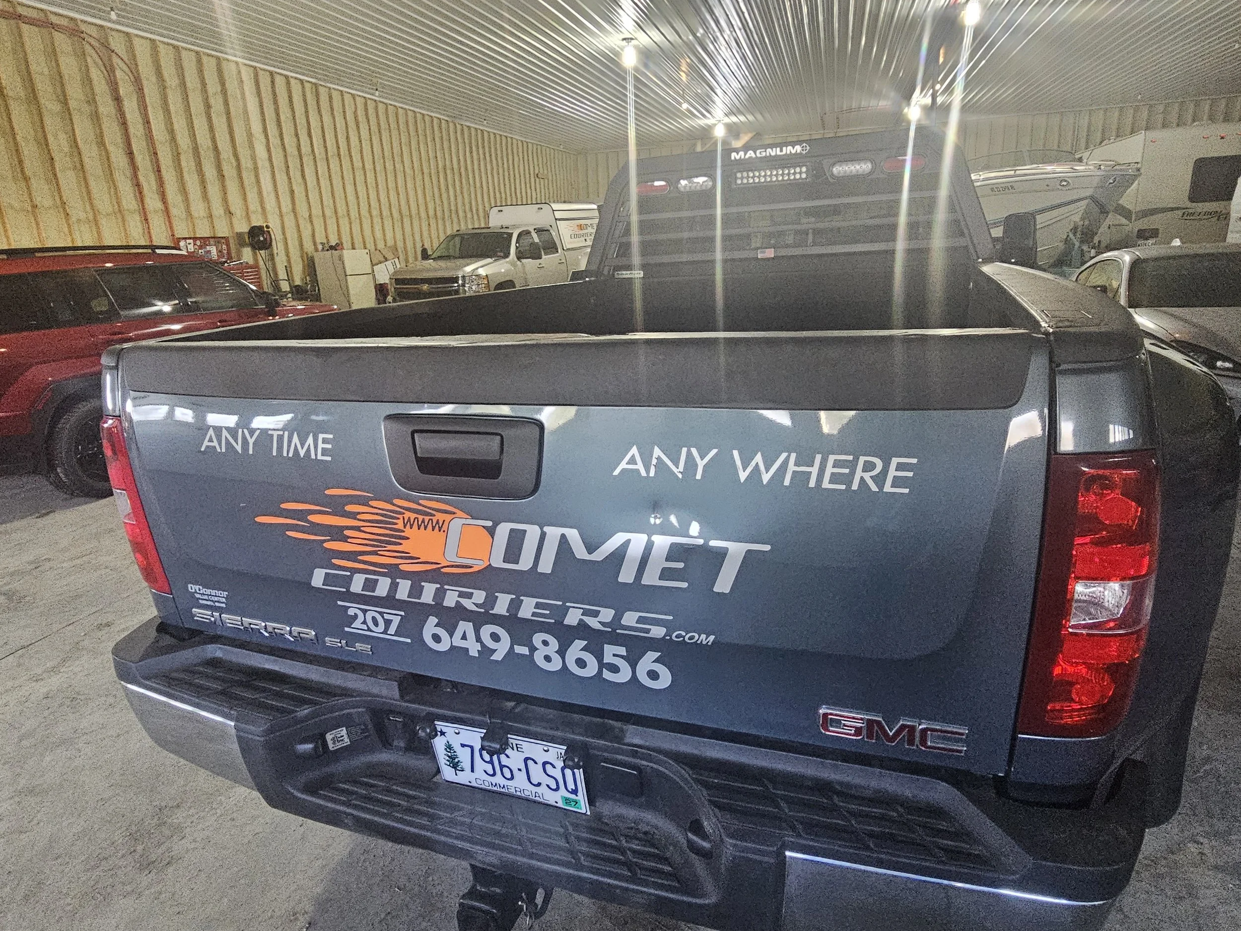 The back of a GMC Sierra pickup truck with advertising stickers for Comet Couriers, including the website and phone number. The truck is inside a warehouse with other vehicles and boats.