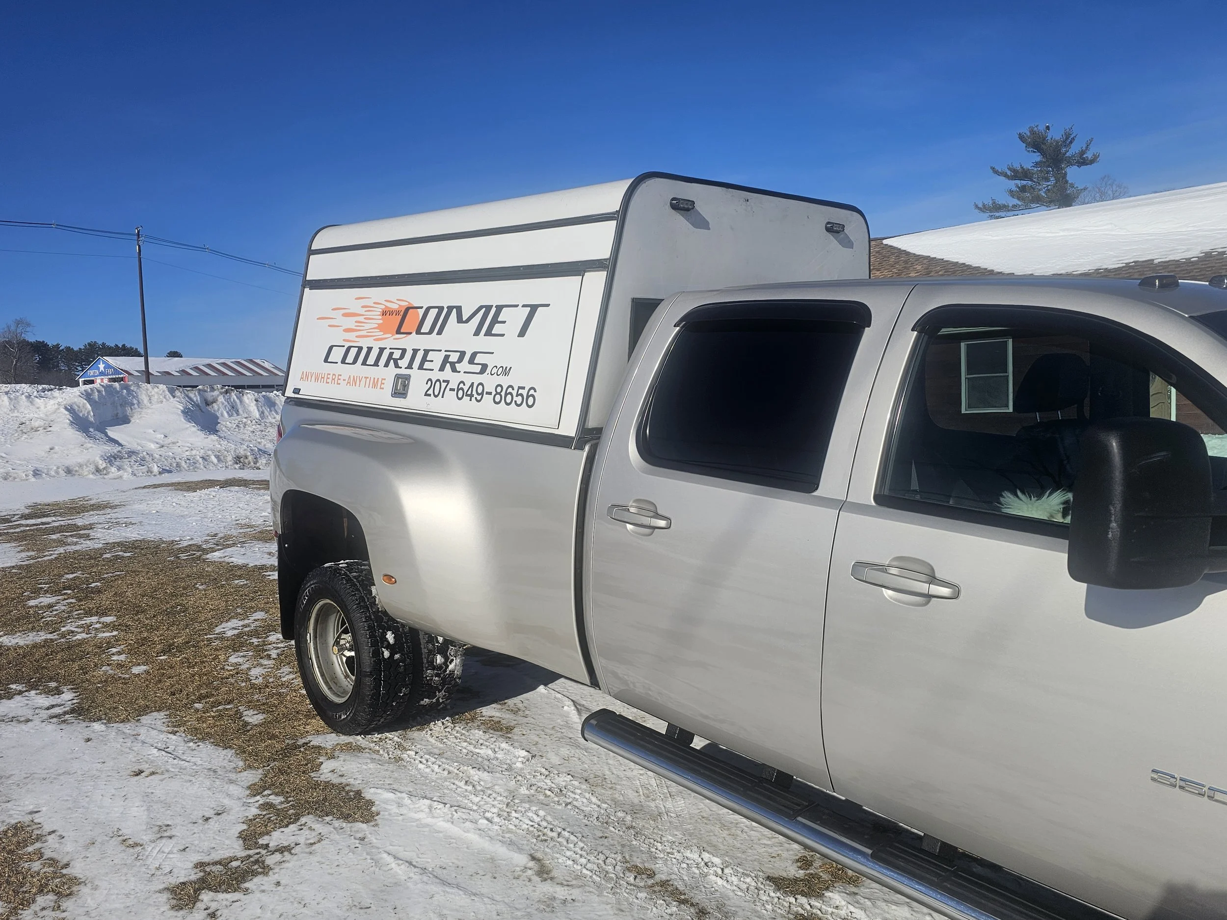 A silver pickup truck with a white Comet Couriers sign on a large box in the truck bed, parked on snow-covered ground on a clear day.