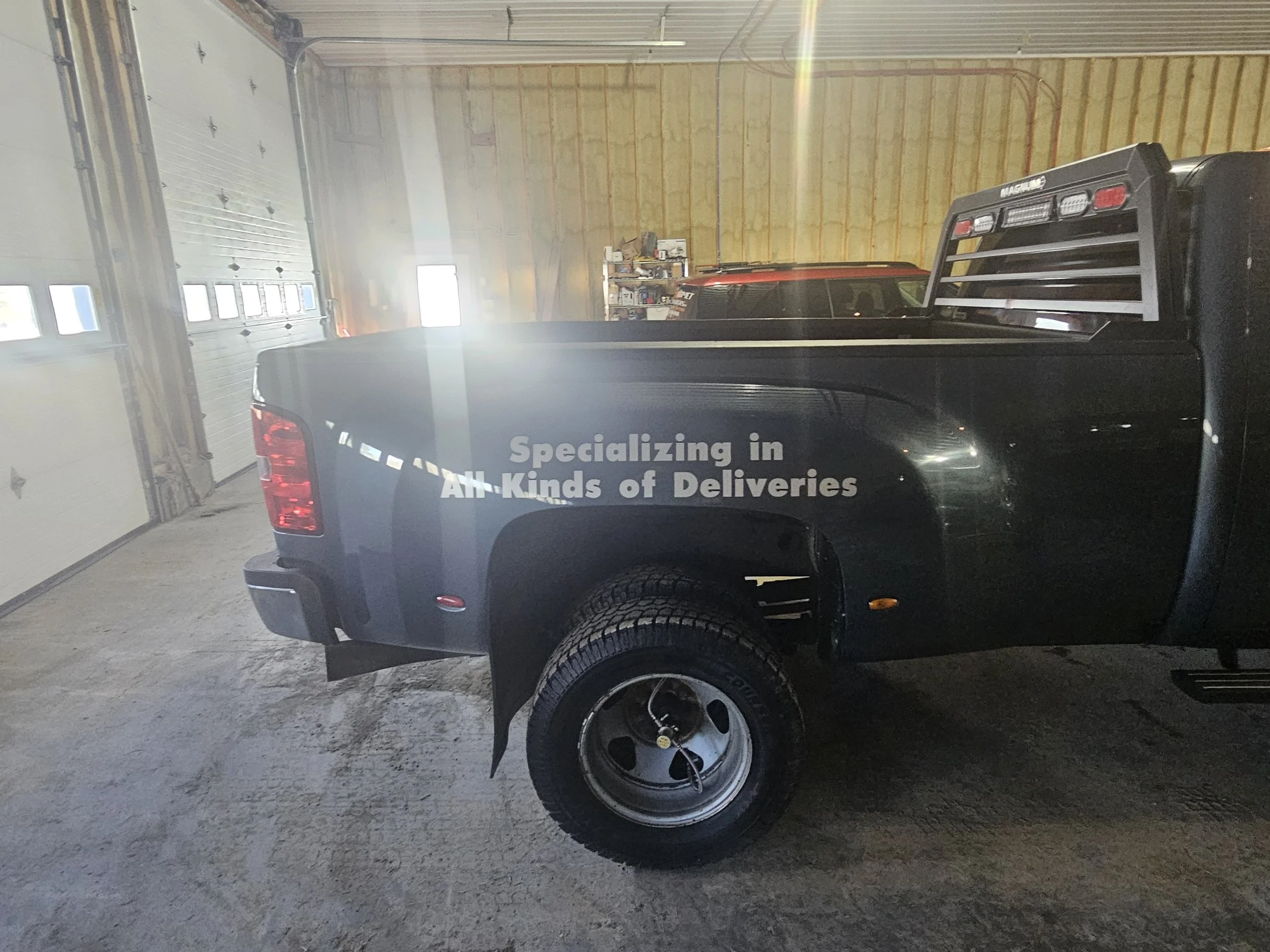 Black pickup truck inside a garage with a partially open garage door. Text on the truck reads 'Specializing in All-Kinds of Deliveries'.